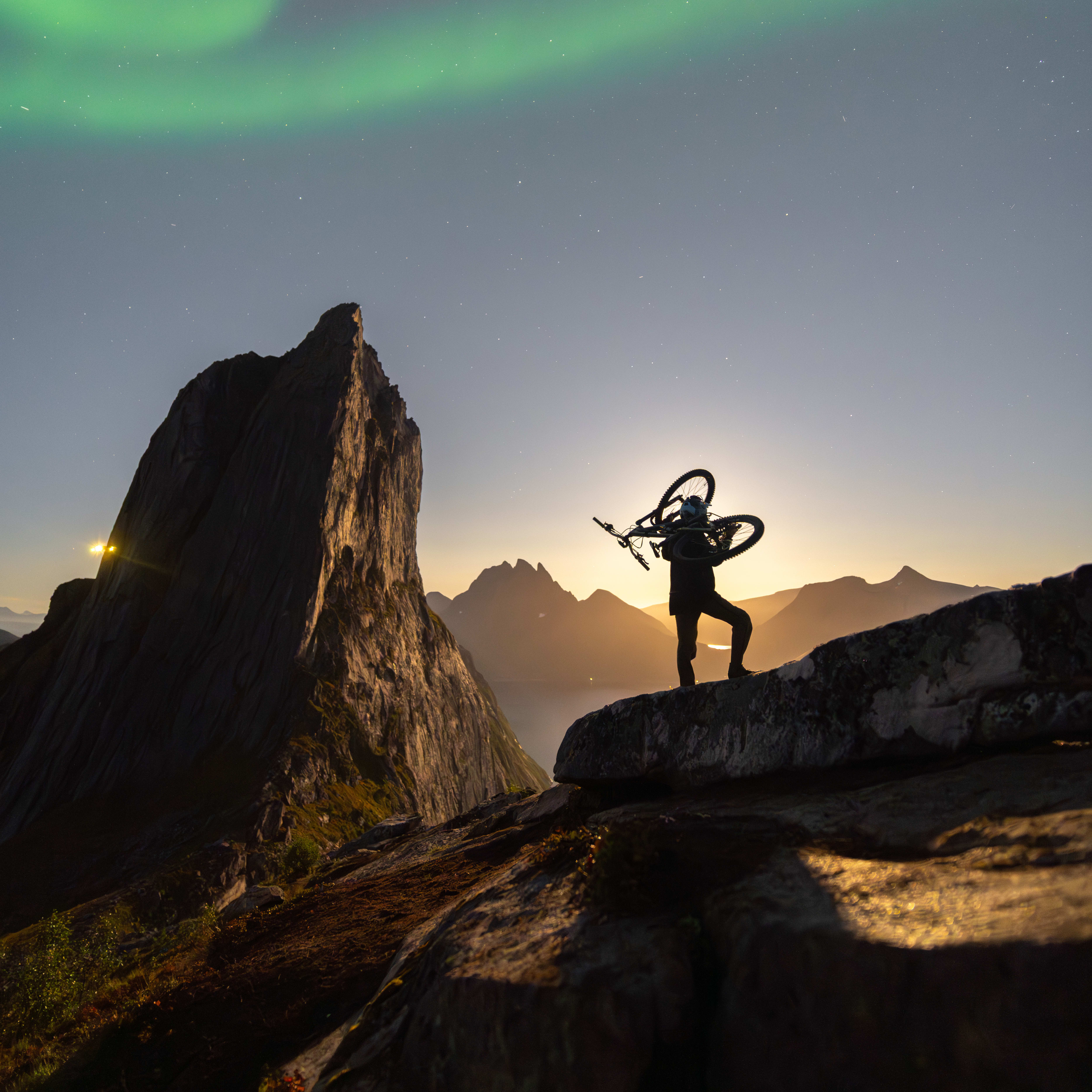 A silhouette of a person with a bike on their shoulders stands on a rocky mountain top with aurora borealis above.