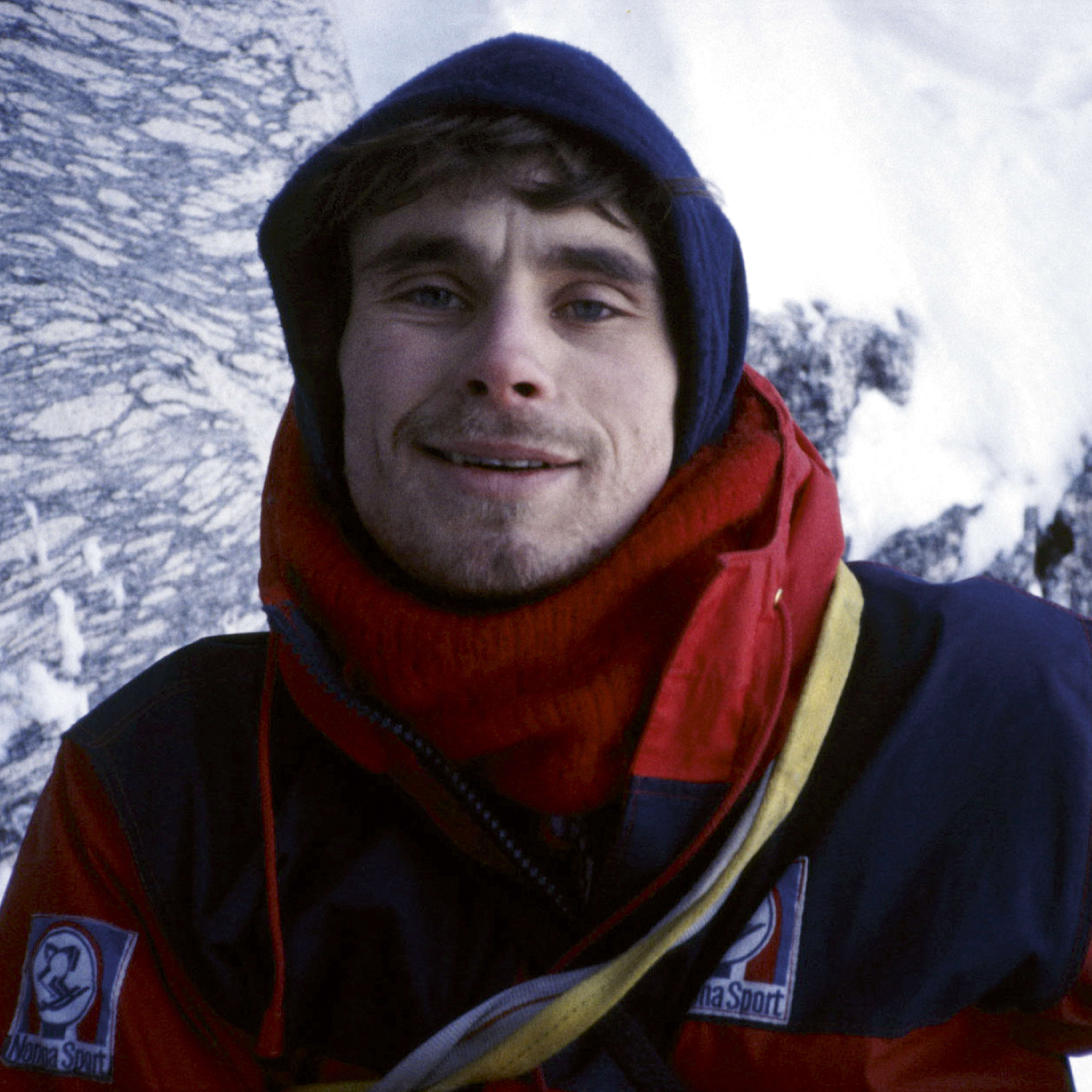 A close-up portrait of a man wearing a blue and red jacket with a white "Nova Sport" logo on the left sleeve and a white logo that says "Nova Sport" on the right sleeve. He is smiling and wearing a red scarf. He is outdoors in a snowy setting.