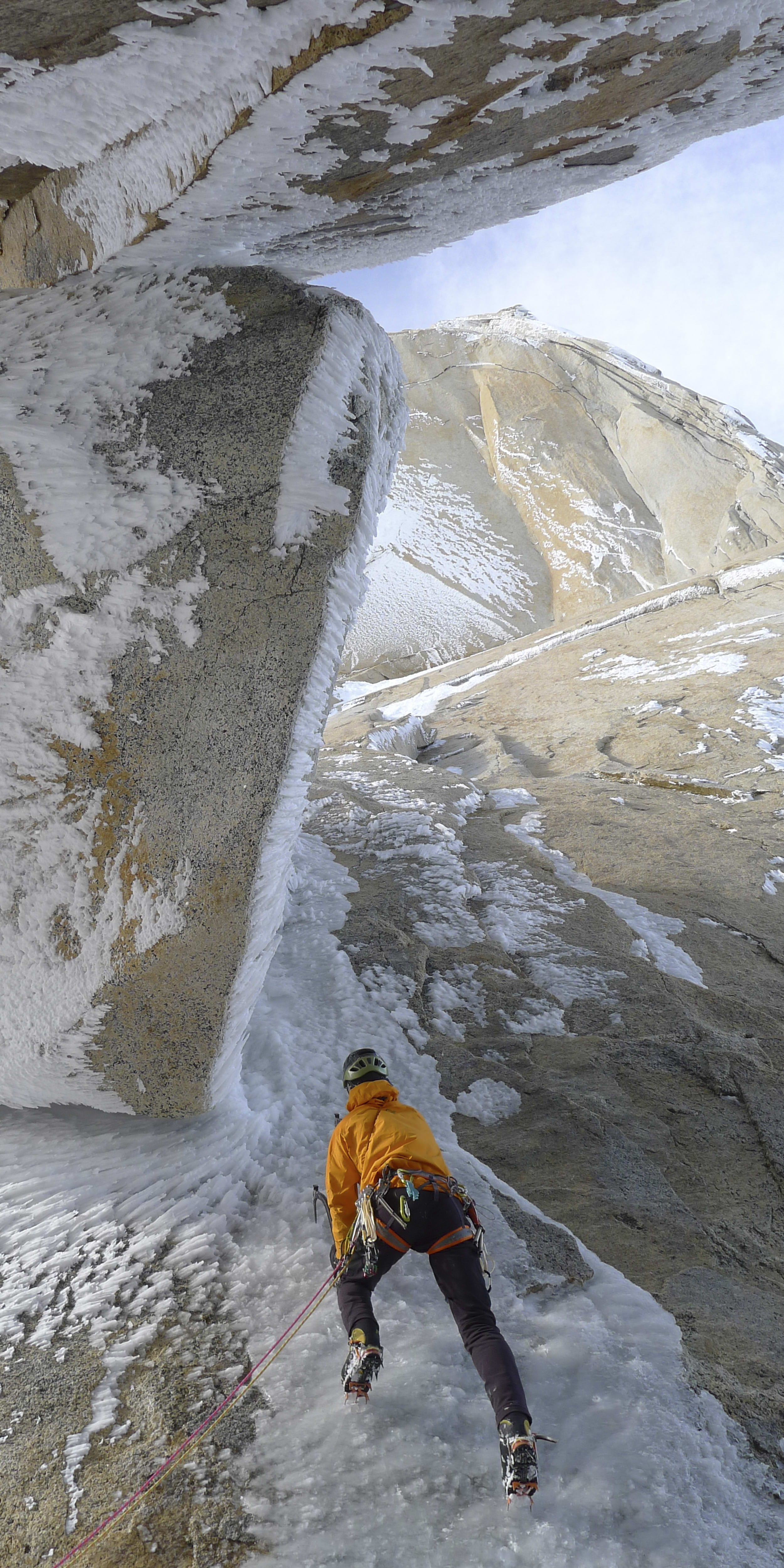 A rock climber in an orange jacket is climbing a snow and ice-covered rock face. The climber is wearing crampons.  The blue sky is visible through the icy overhang. 
