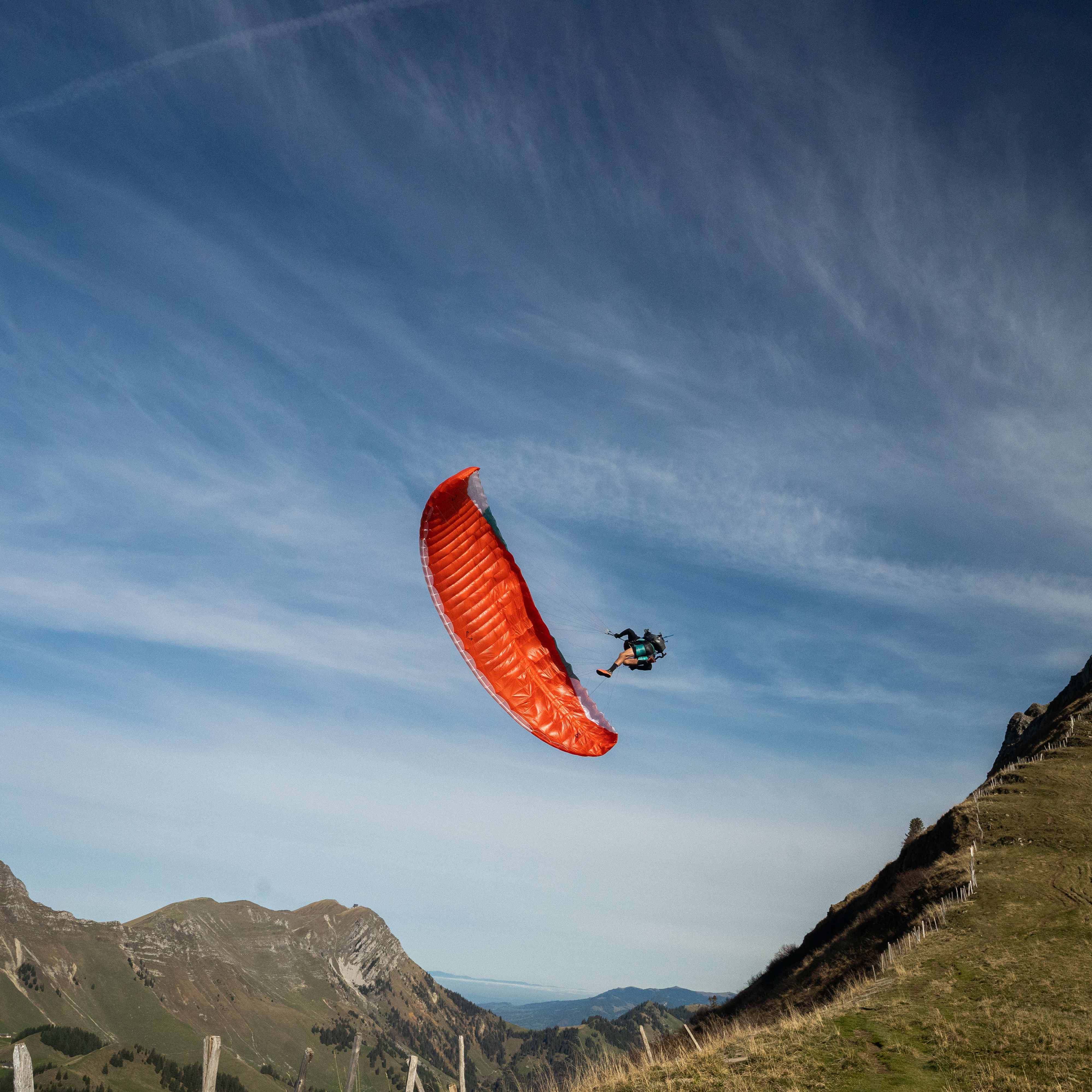 A red paraglider flies over a grassy hillside with a mountain in the background. The sky is blue and clear, with a few wispy clouds.  
