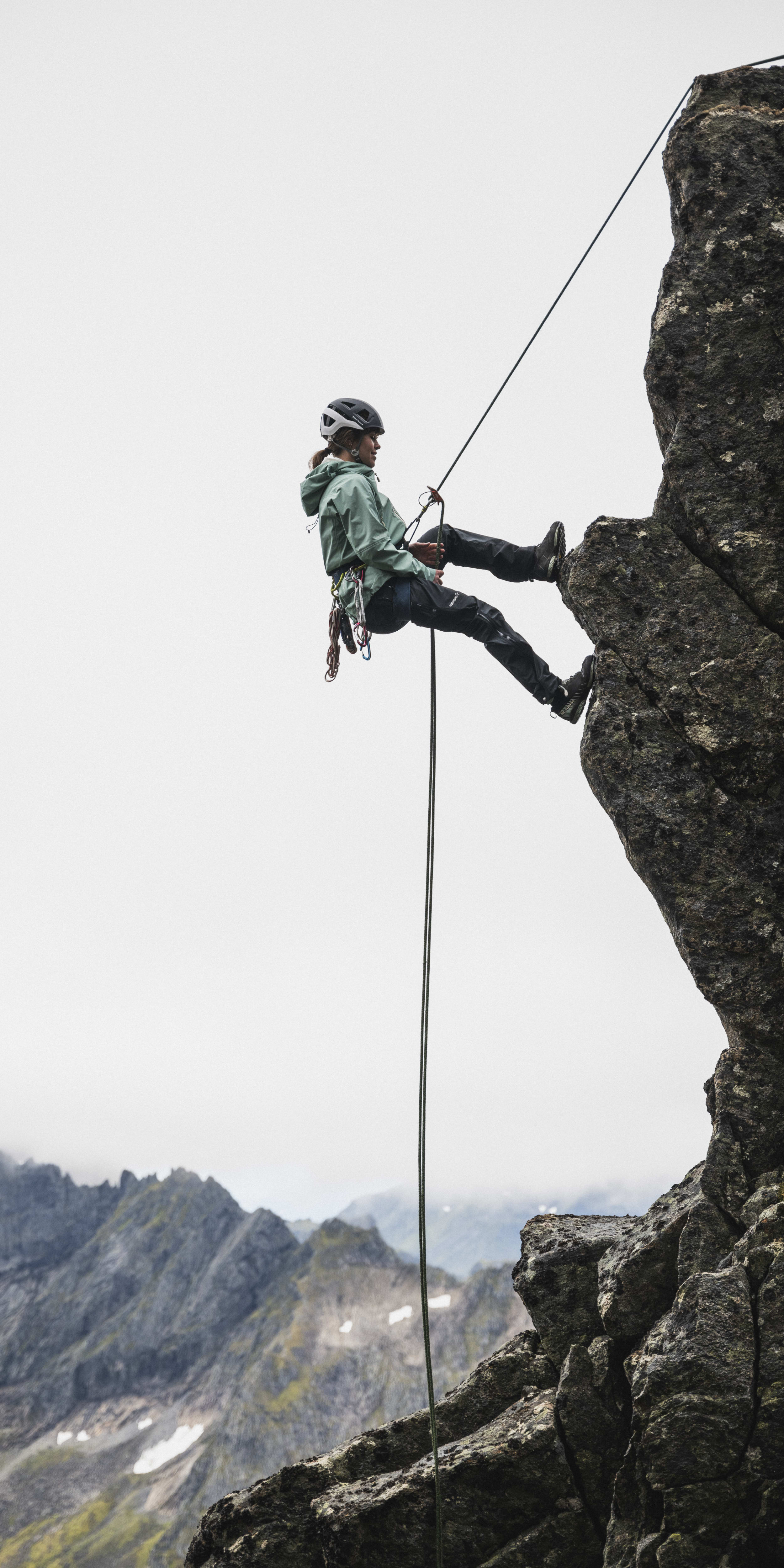 A climber wearing a helmet and Norrøna clothing rappels down a rocky cliff.