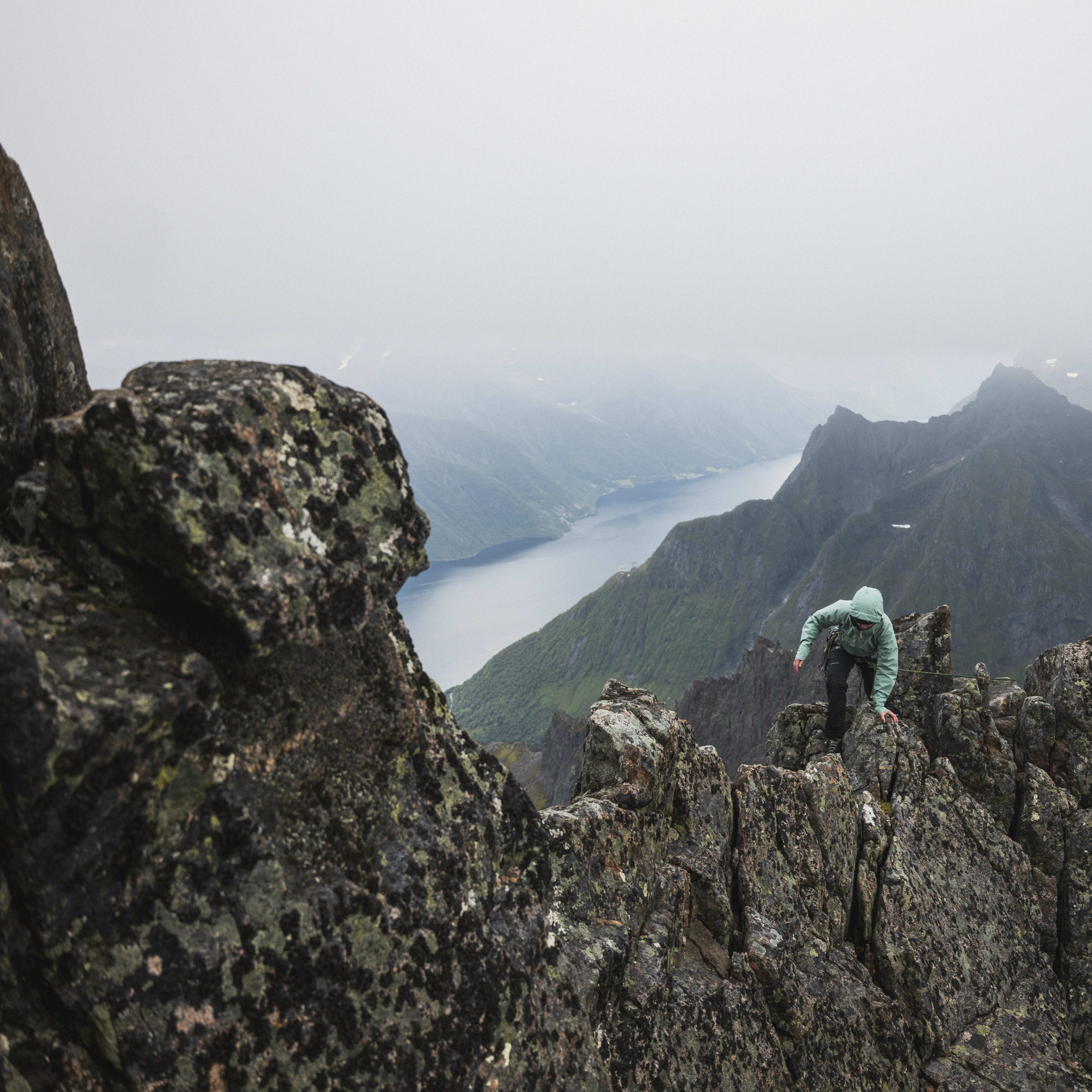 A person wearing Norrøna clothing climbs a rocky mountain ridge. The view shows a fjord in the distance.