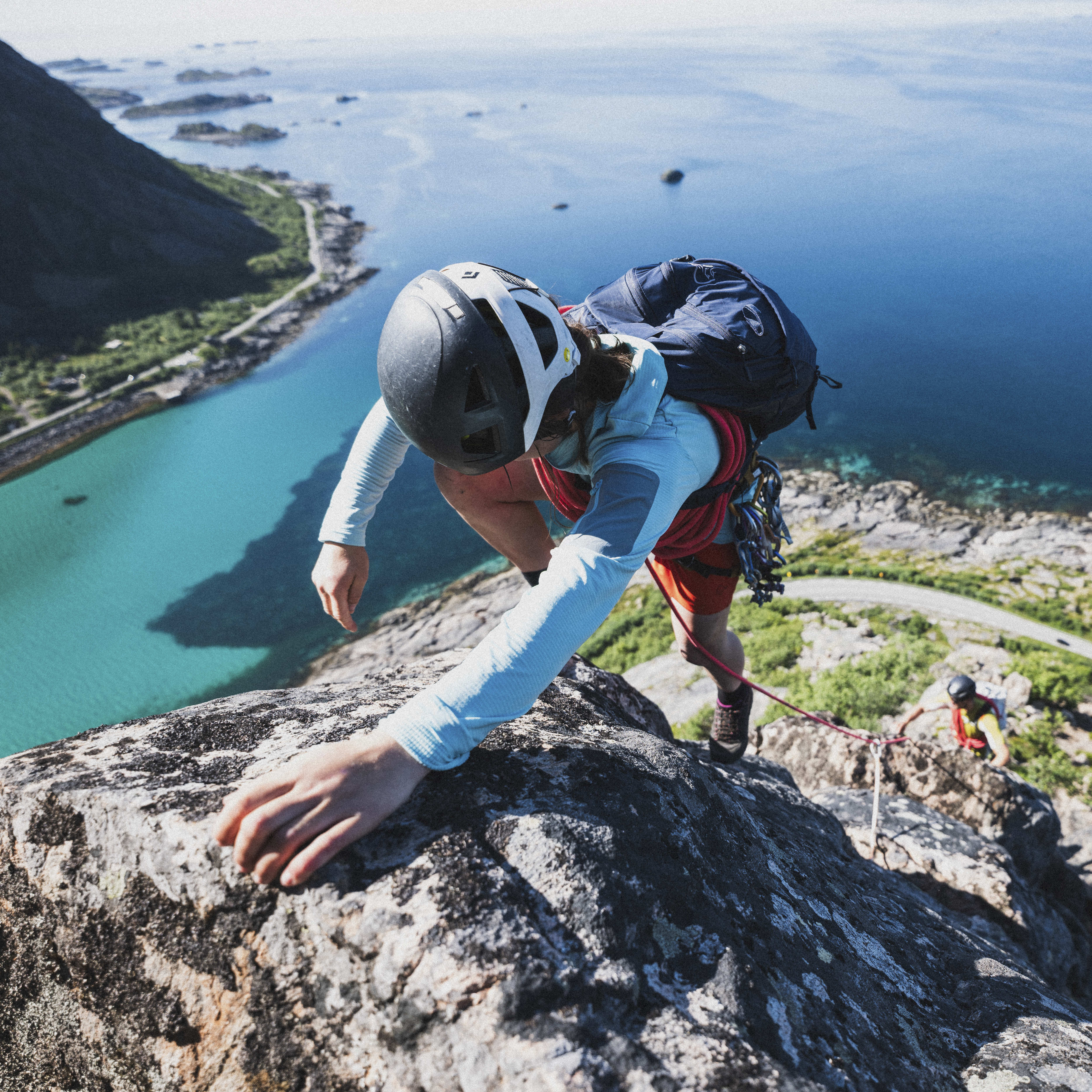 A climber scales a rocky cliff face with turquoise water below. They wear a helmet, backpack, and a Norrøna light-blue jacket. Another climber is visible further down.
