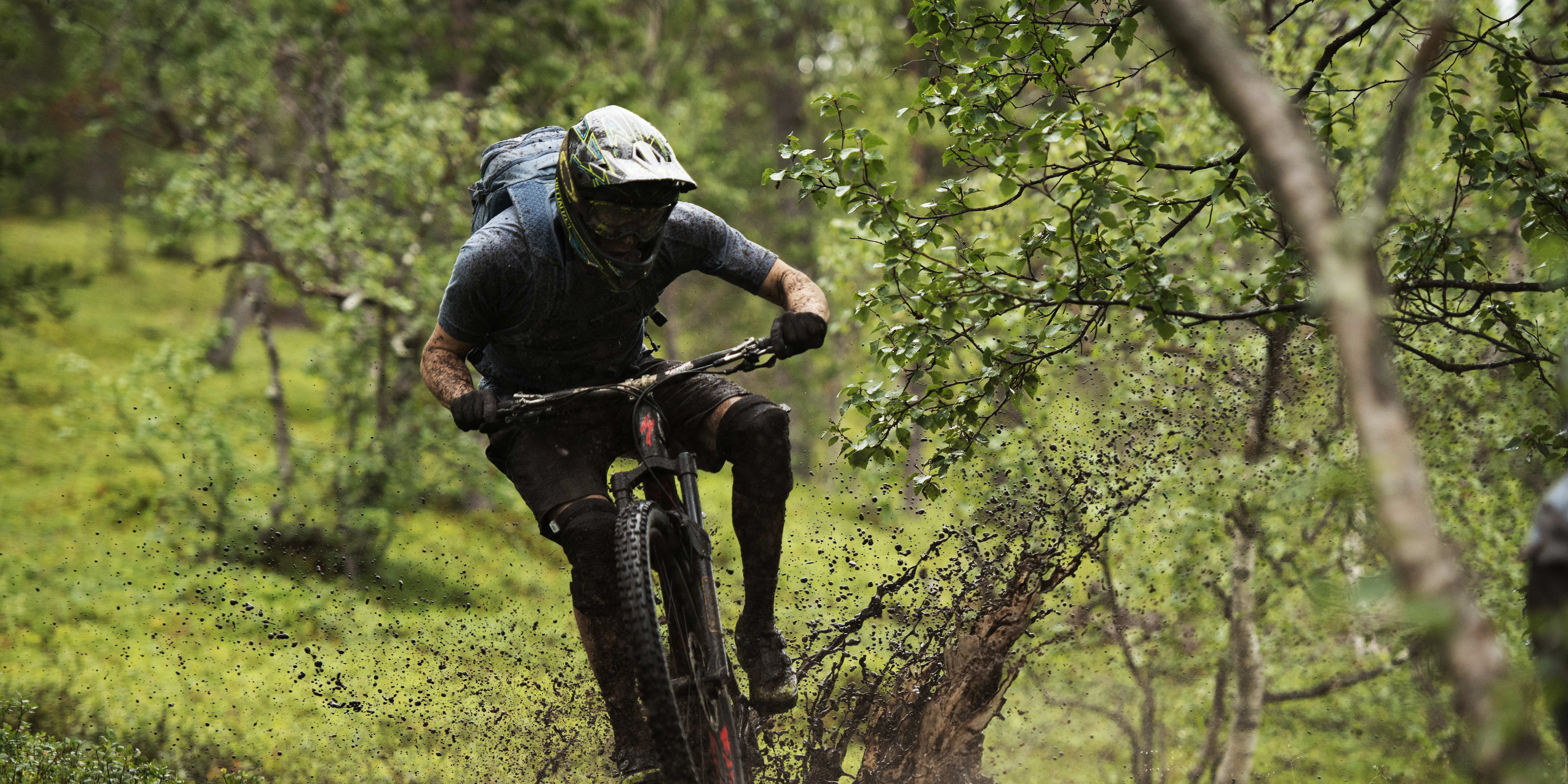 A person in a Norrøna cycling outfit rides a mountain bike through a muddy forest path, kicking up a spray of dirt.