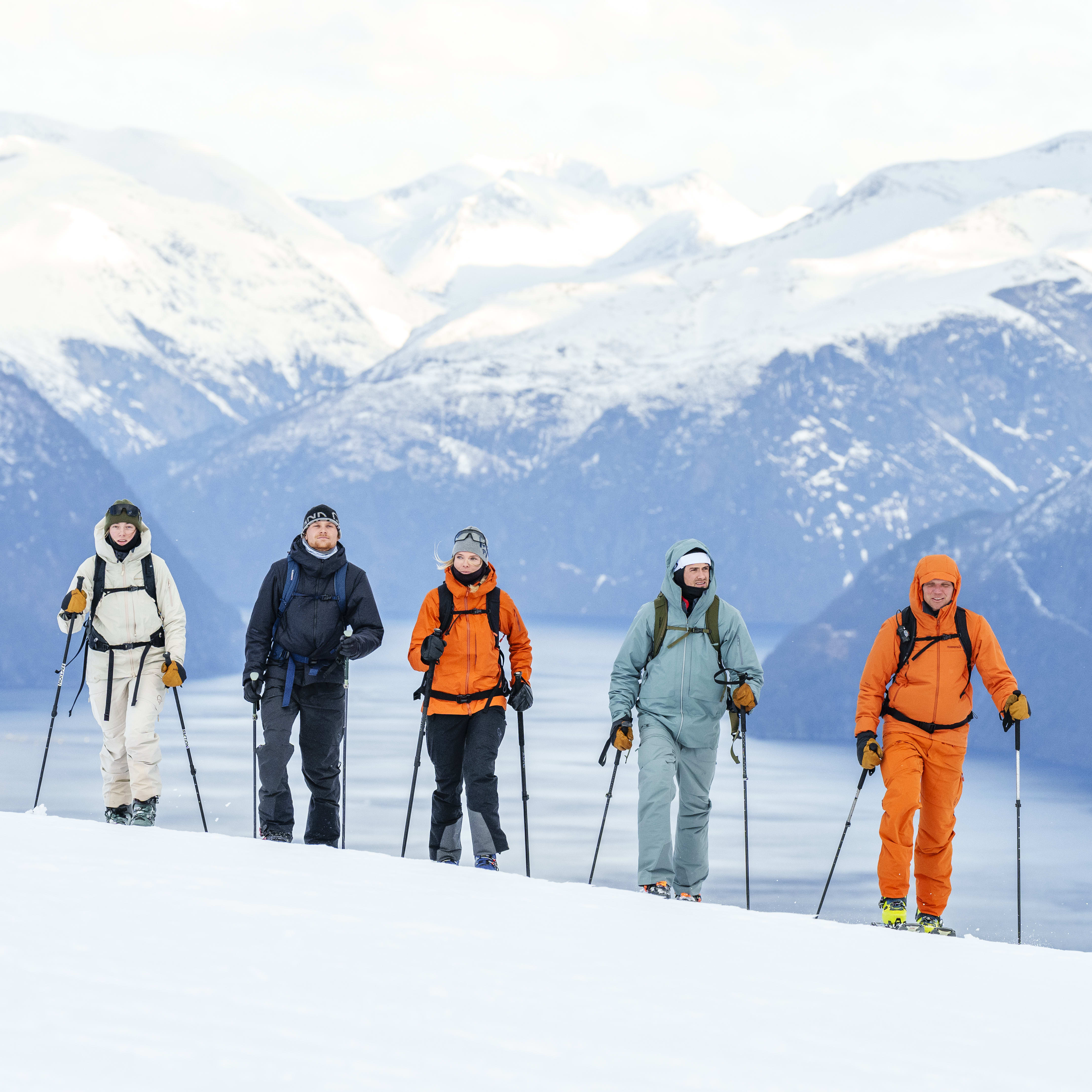 Five people wearing Norrøna clothing are backcountry skiing in a snowy mountain landscape. 
