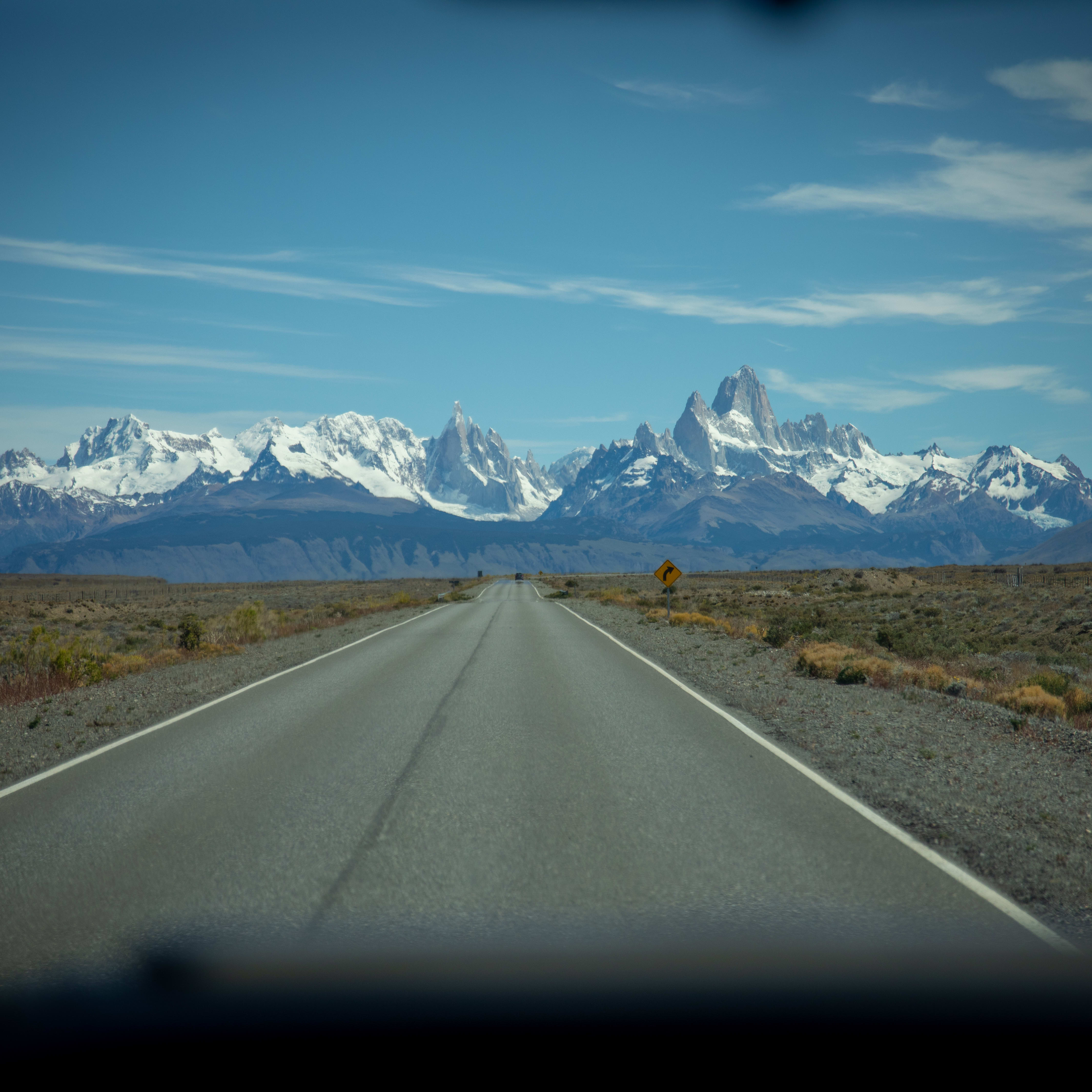 A paved road with a white dividing line leading towards snow-capped mountains. The road is flanked by brown brush and a yellow road sign. 

