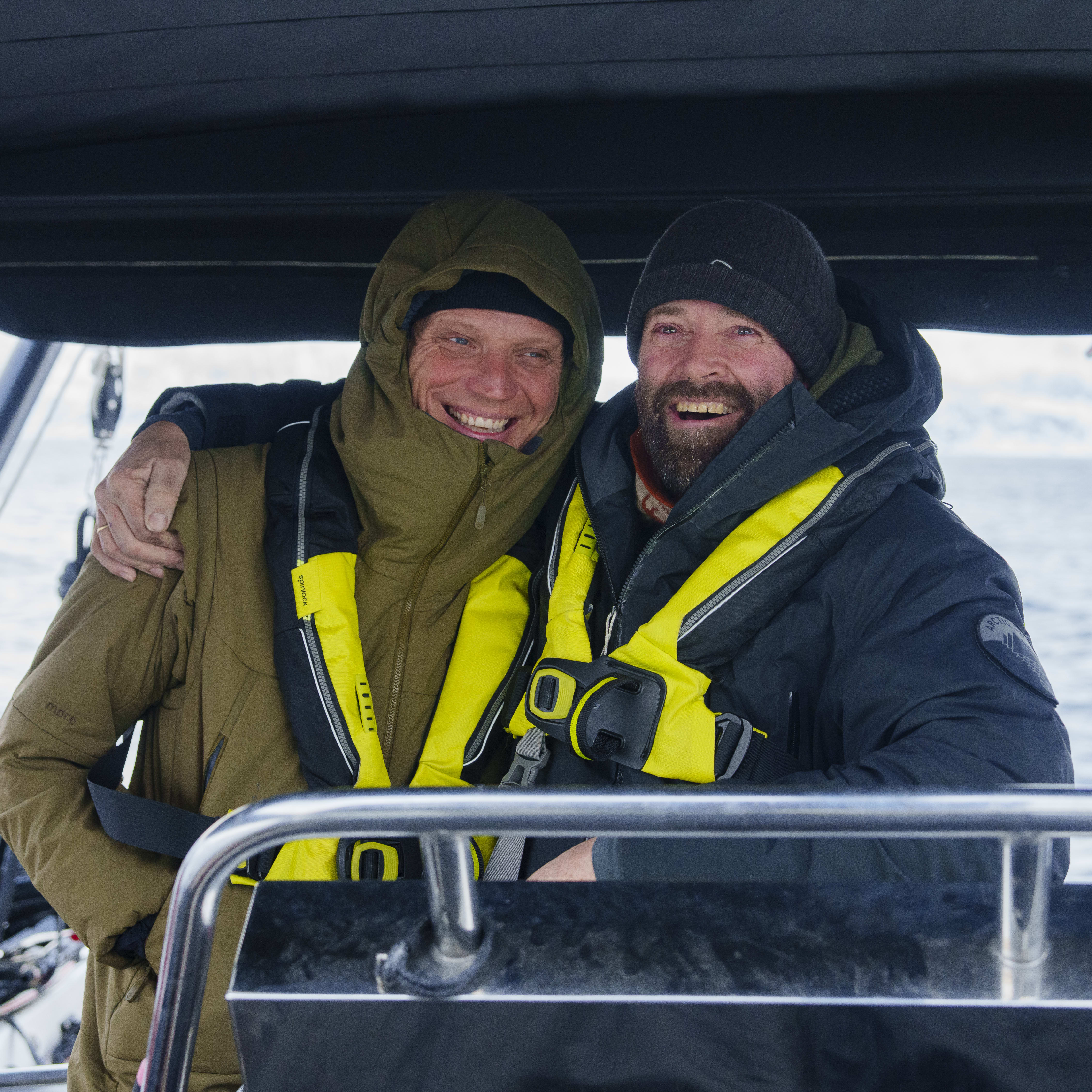 Two smiling men on a boat wearing Norrøna jackets and life vests. Water and mountains are in the background.