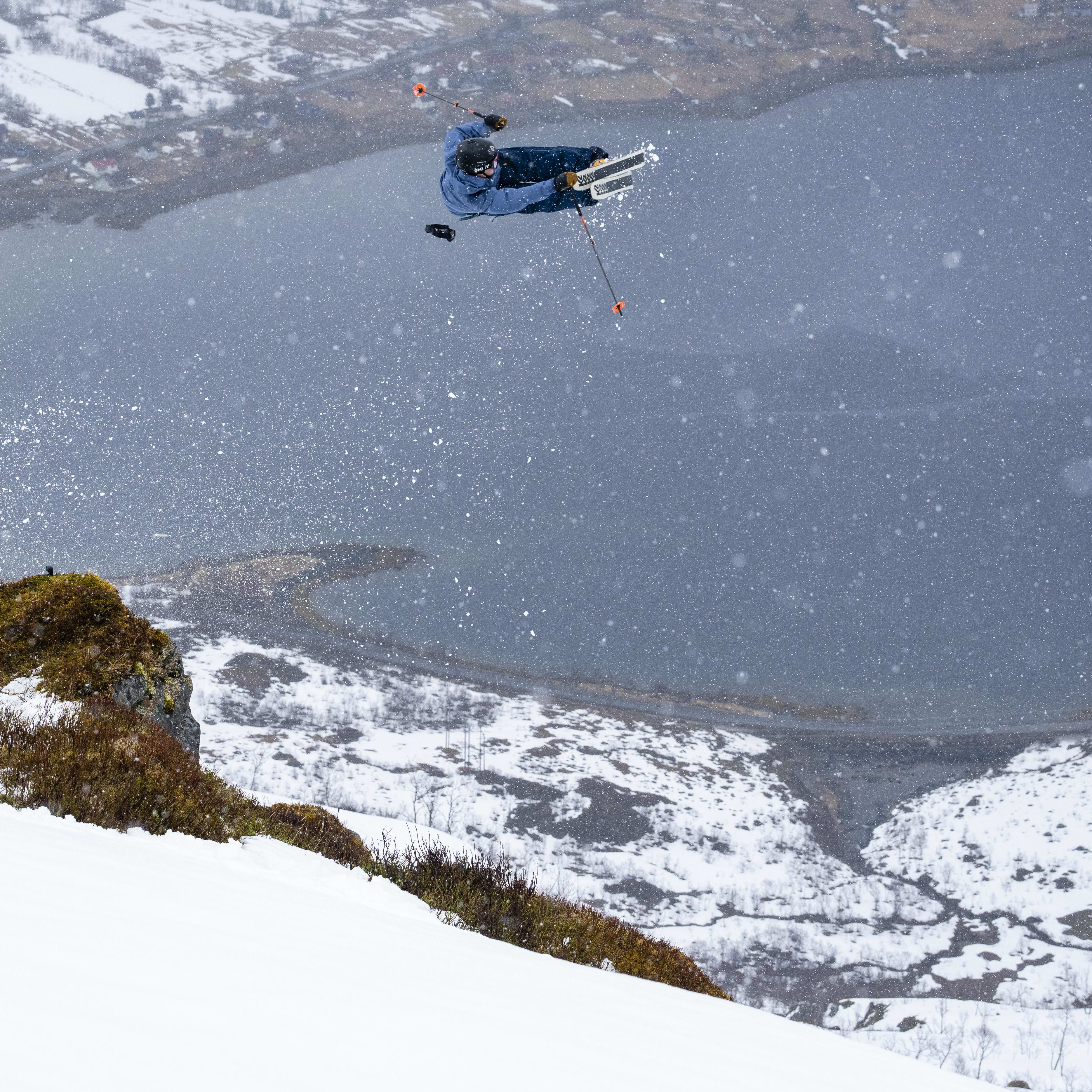 A skier in a blue jacket and helmet performs a trick while skiing off a snow-covered mountain. It is snowing and the background includes a frozen lake and snowy mountains.