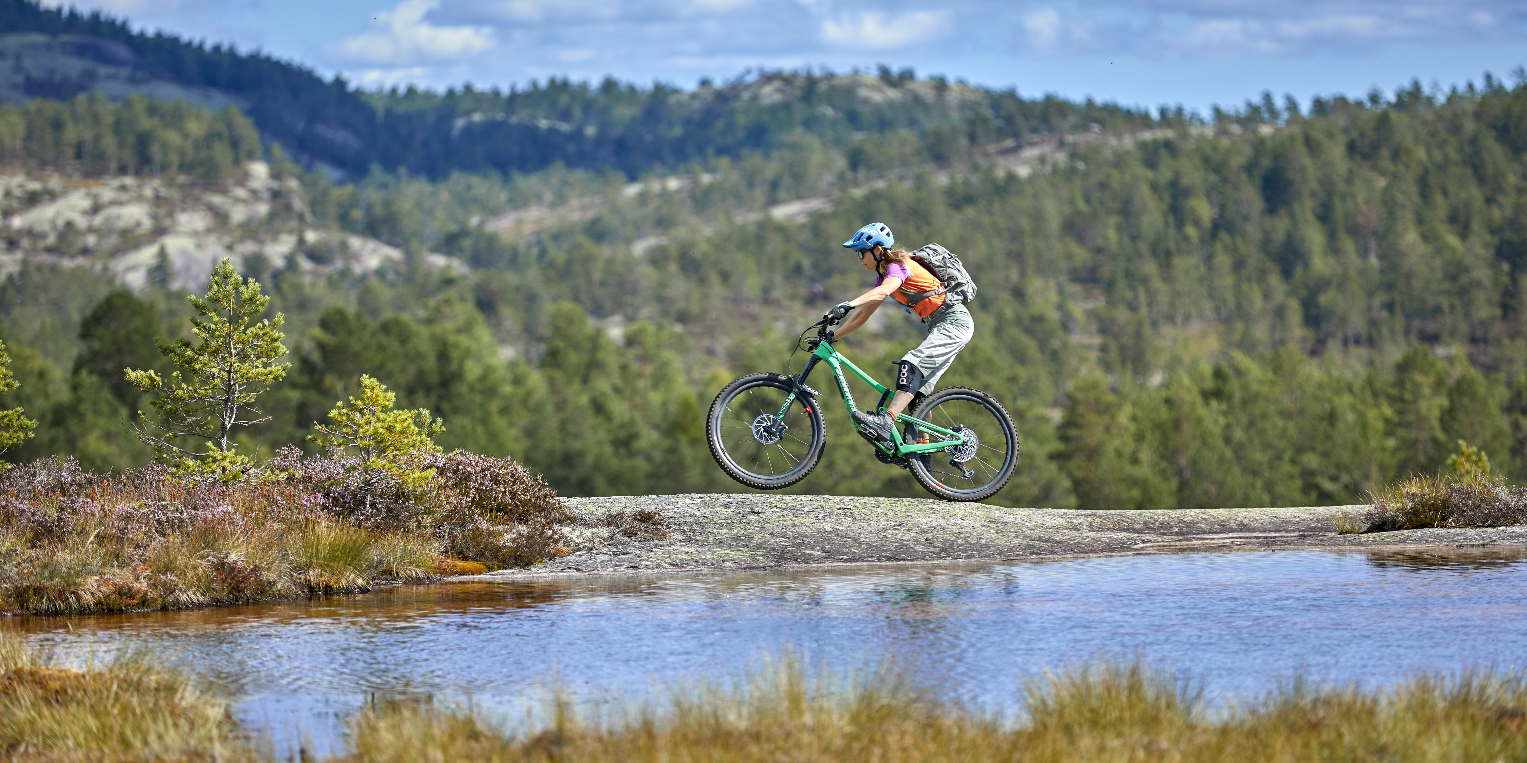 A person in Norrøna clothing mountain biking on a rock formation with a backdrop of green hills and a blue sky with clouds.