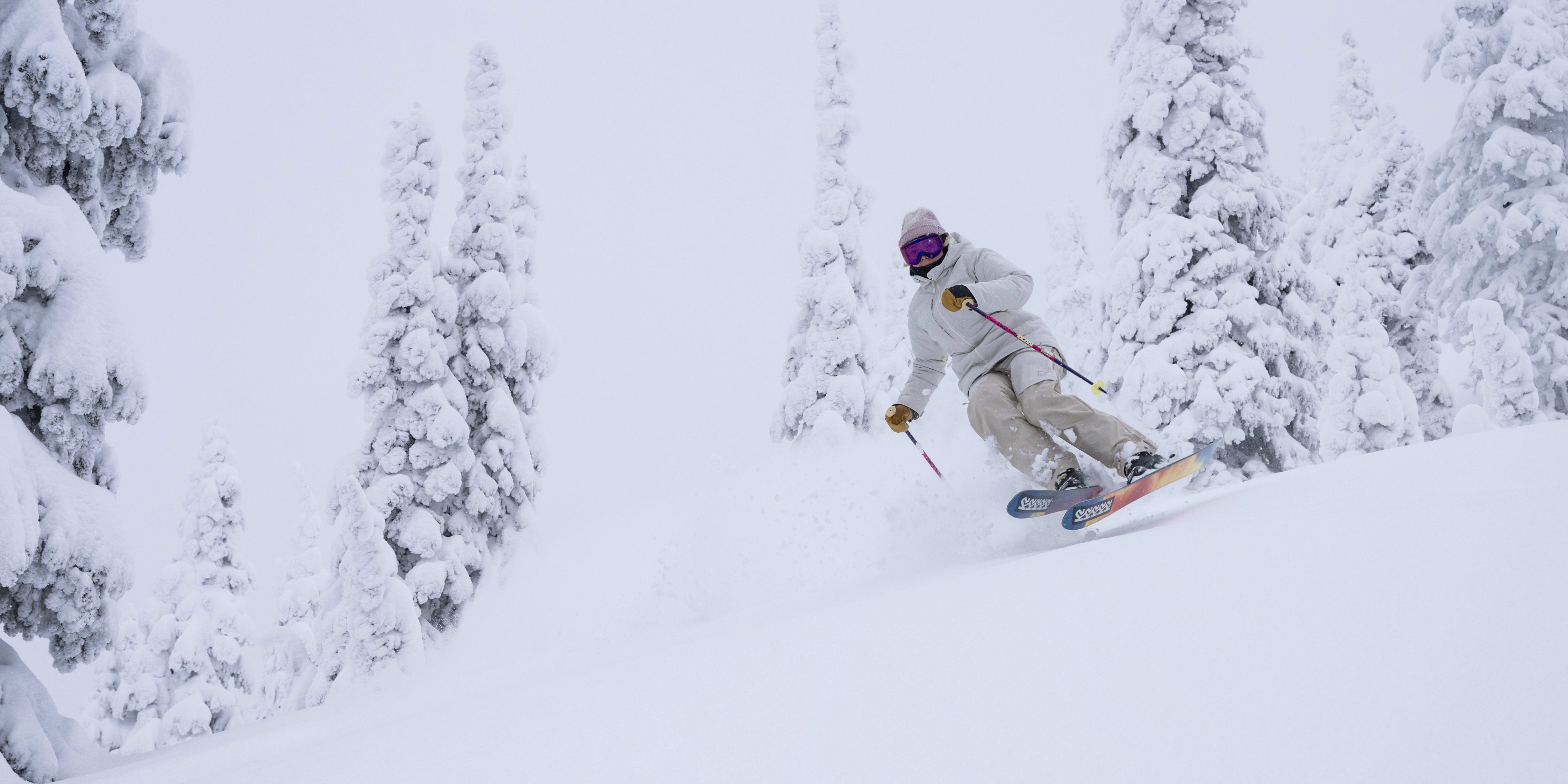 A person wearing Norrøna clothing skis down a snowy slope. Trees are covered in snow in the background.