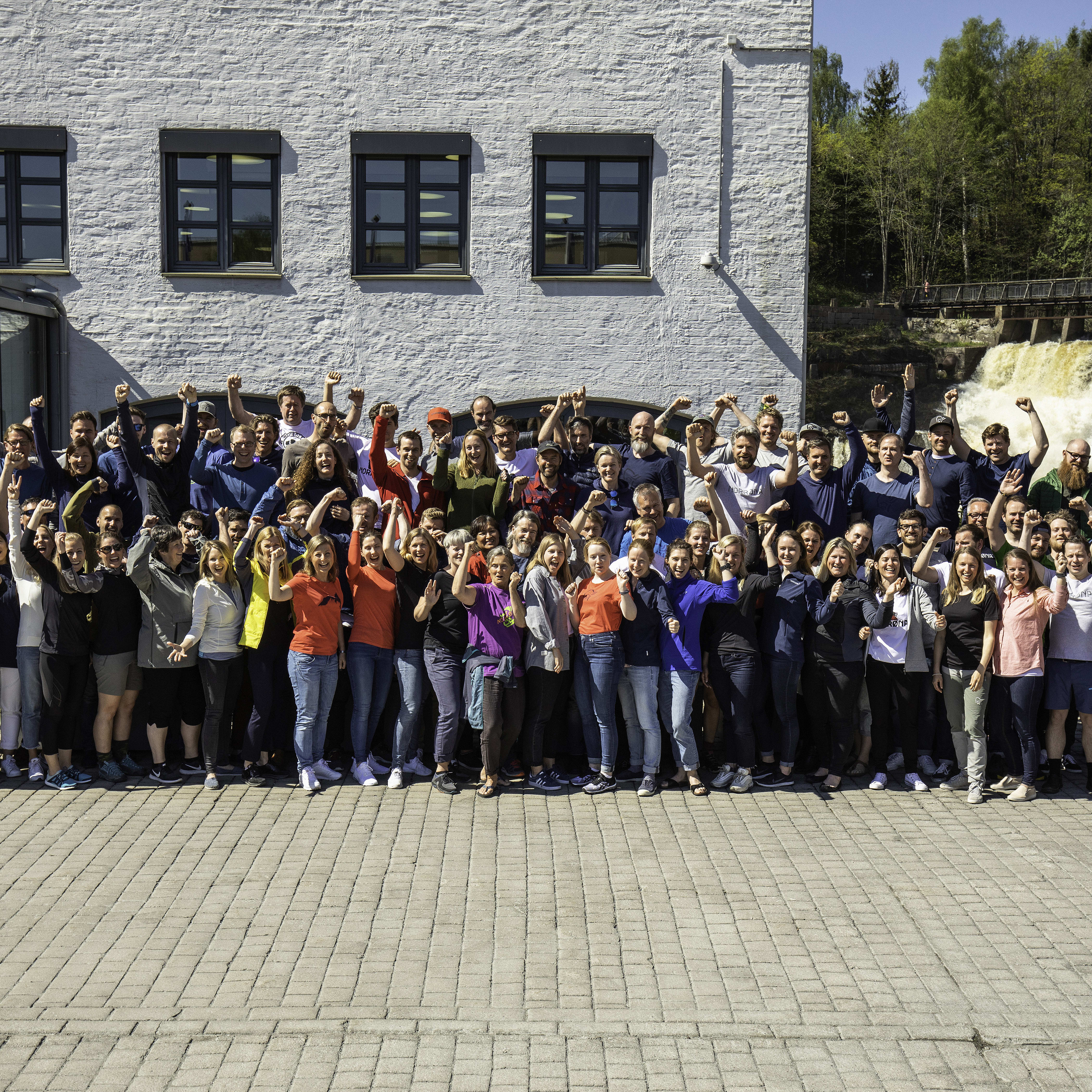 A large group of people stand outside of the Norrøna headquarters cheering with their arms raised. A waterfall is behind them.