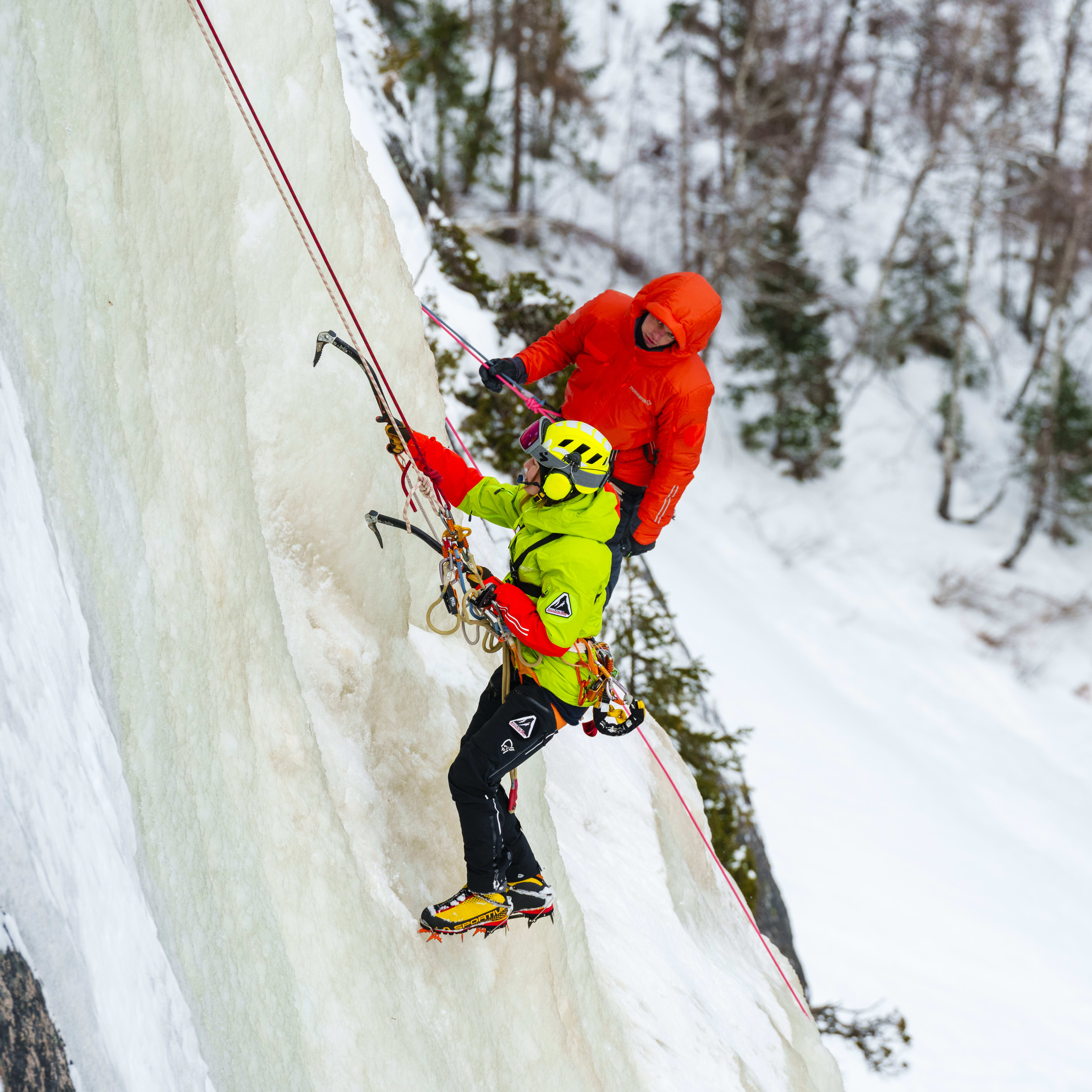 Two climbers, wearing Norrøna gear, ice climbing on a snowy cliff face. The climber on the left is belaying the climber on the right.