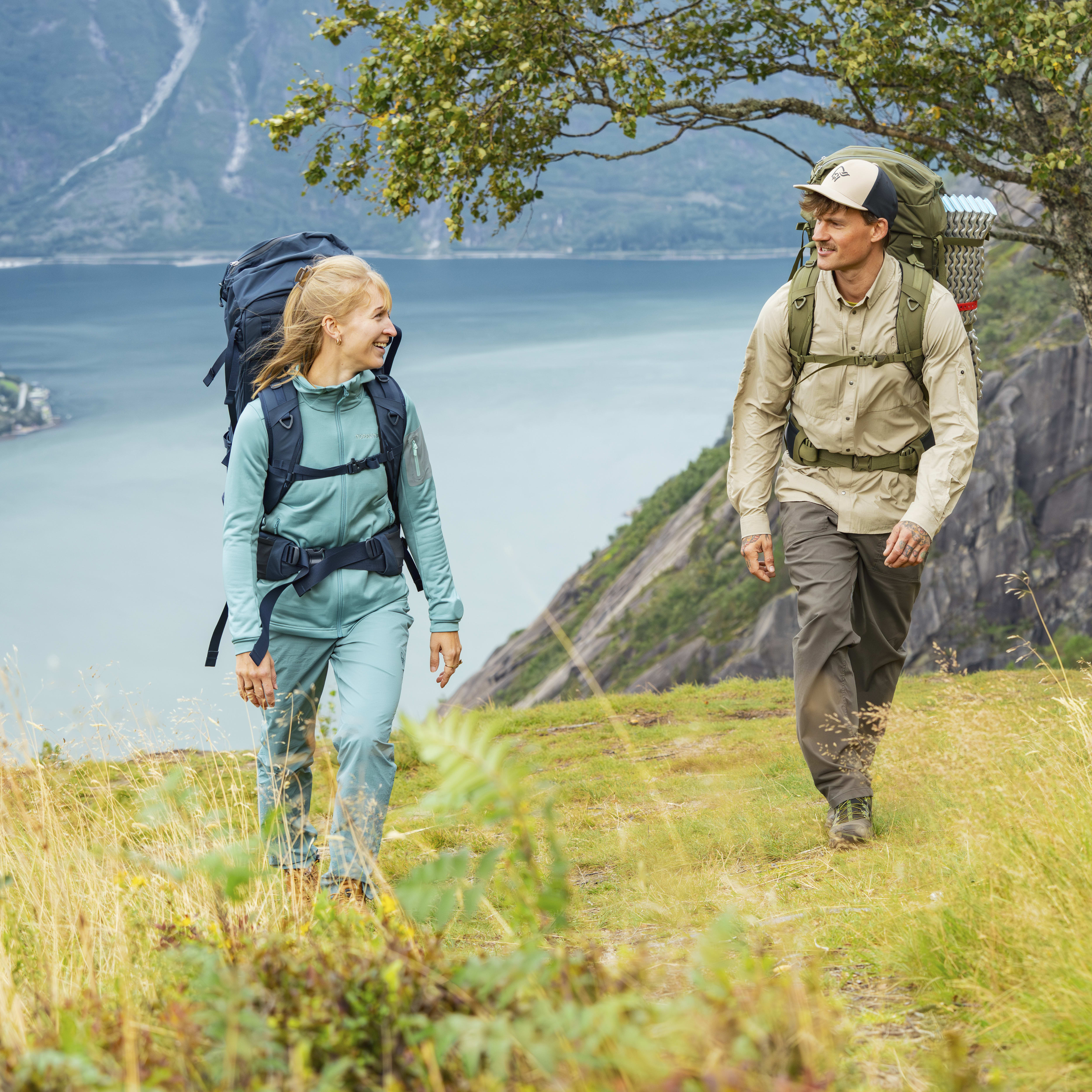 A smiling couple hikes on a grassy trail overlooking a fjord. The woman wears a blue Norrøna fleece jacket and the man wears a tan Norrøna shirt.
