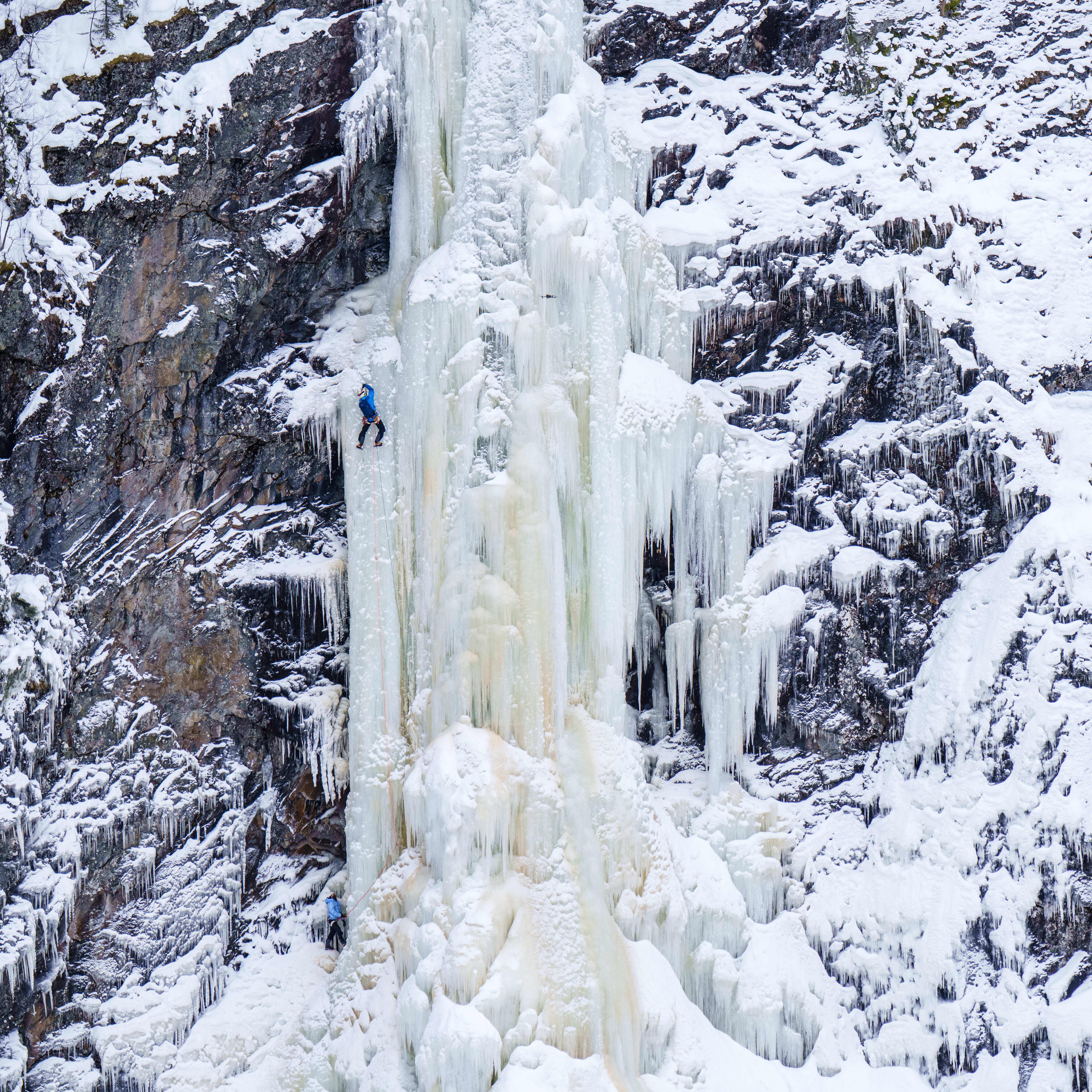 Two ice climbers wearing Norrøna clothing scale a massive, frozen waterfall with snowy rock faces.