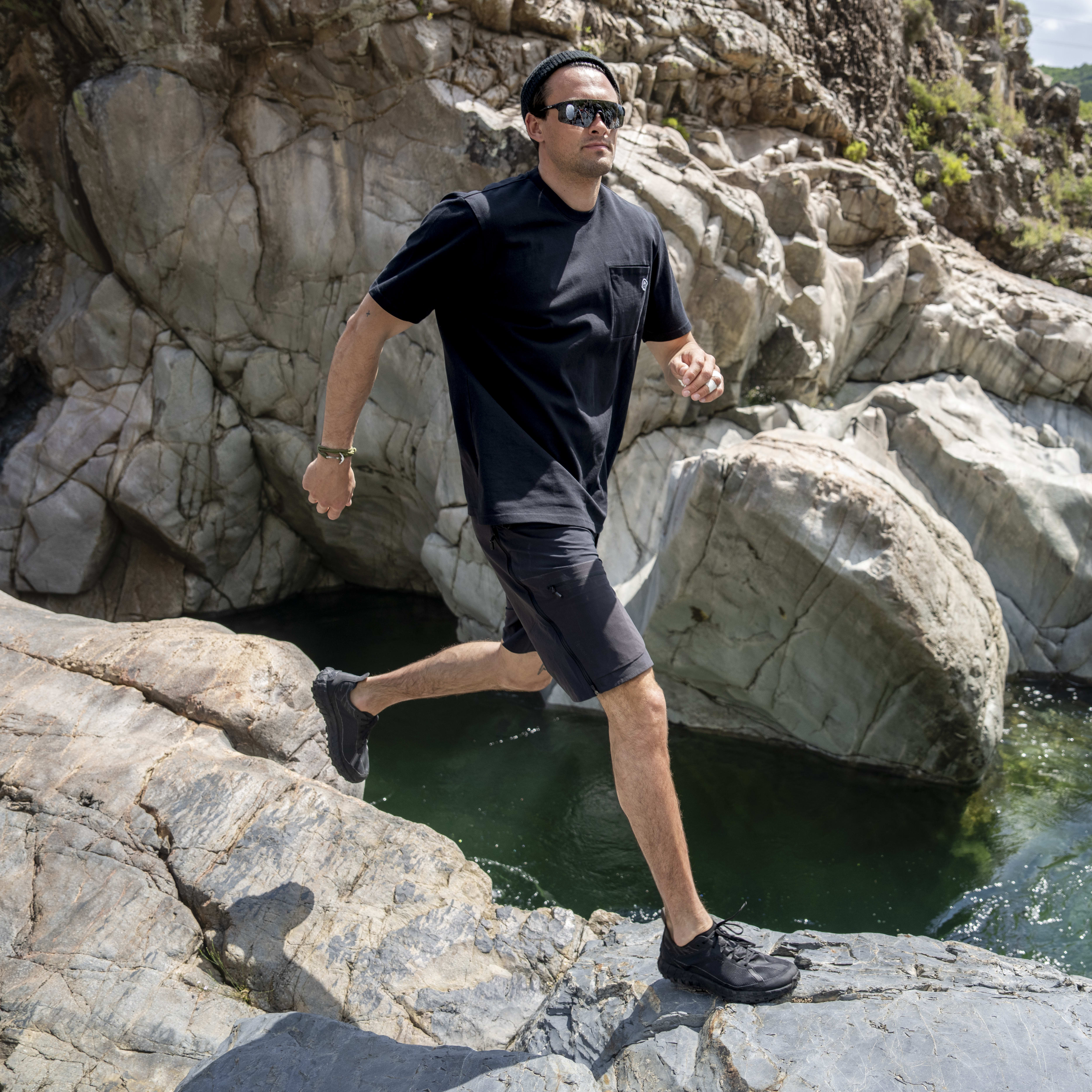 Man in black Norrøna shorts jumps across rocks near a river.  
