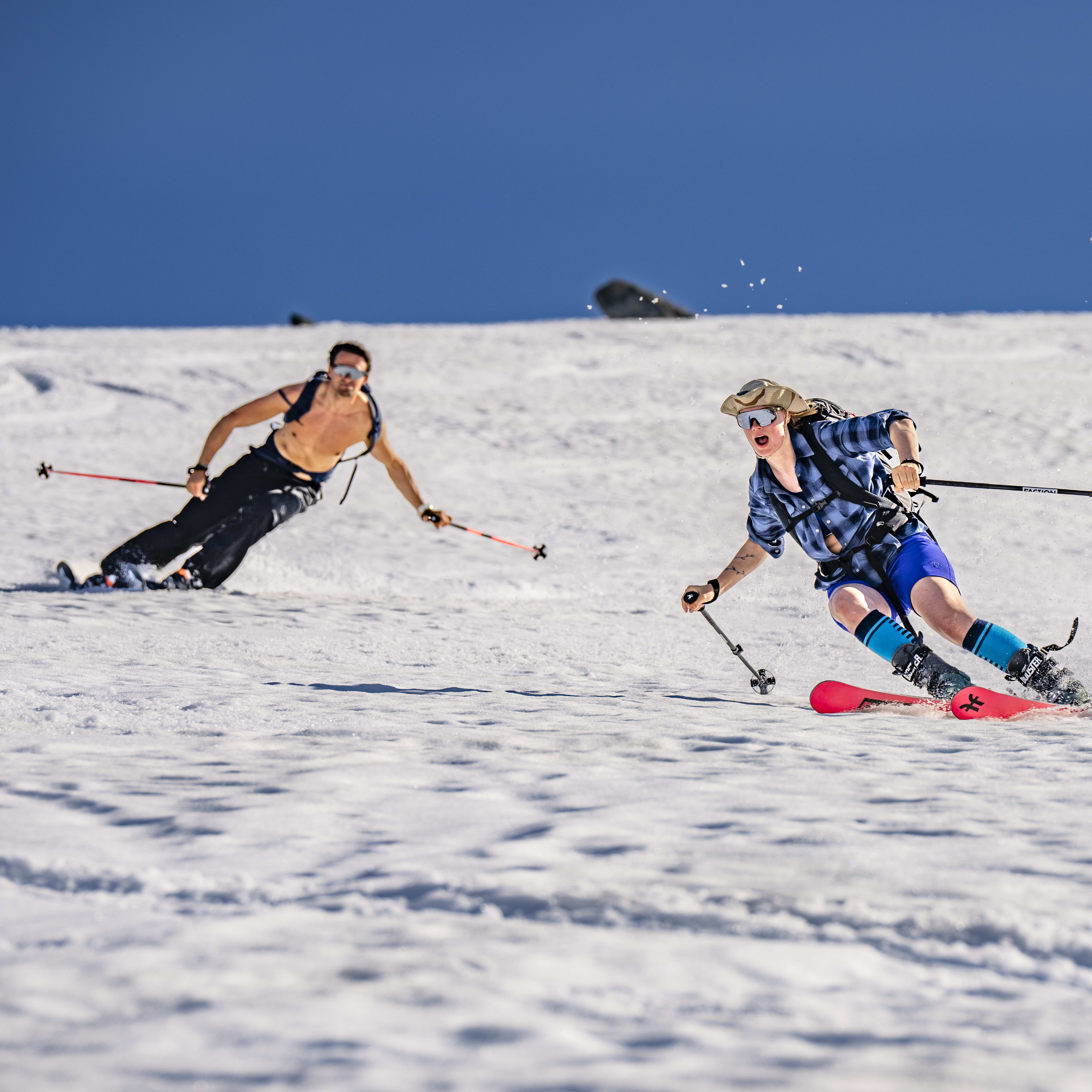 Two people ski down a snowy slope, a person in a blue plaid shirt is in the foreground, and a person in a black shirt is in the background.  Both skiers are holding poles.  The sky is blue.