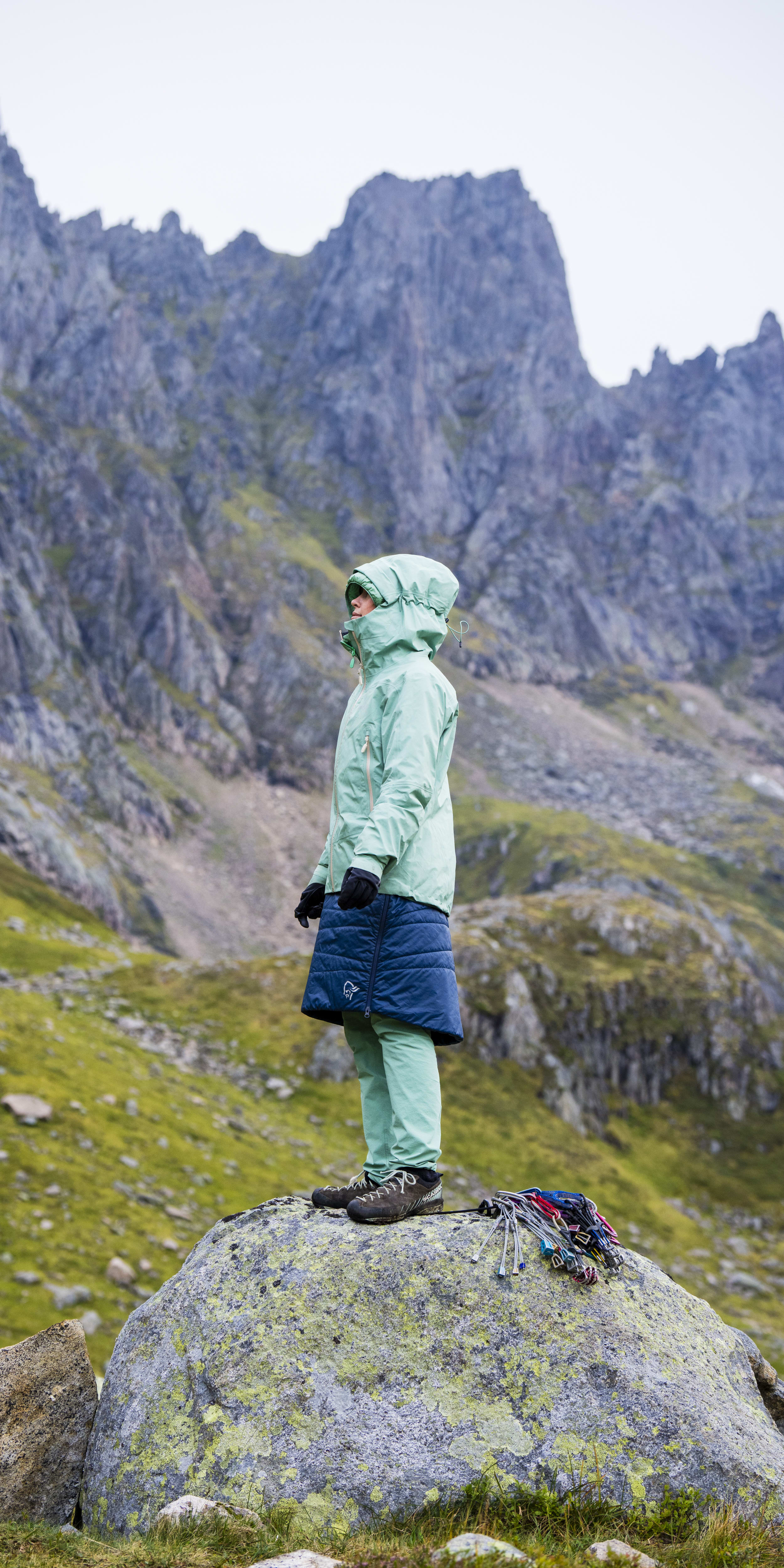 A woman wearing a Norrøna jacket stands on a rock in front of a mountain range. She is wearing a Norrøna skirt and has climbing gear on the rock next to her. 
