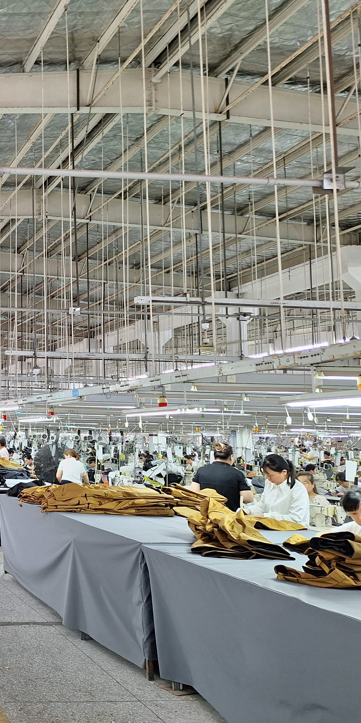 A large factory room with many people working at sewing machines.  There is a large number of Norrøna clothing items on tables. 
