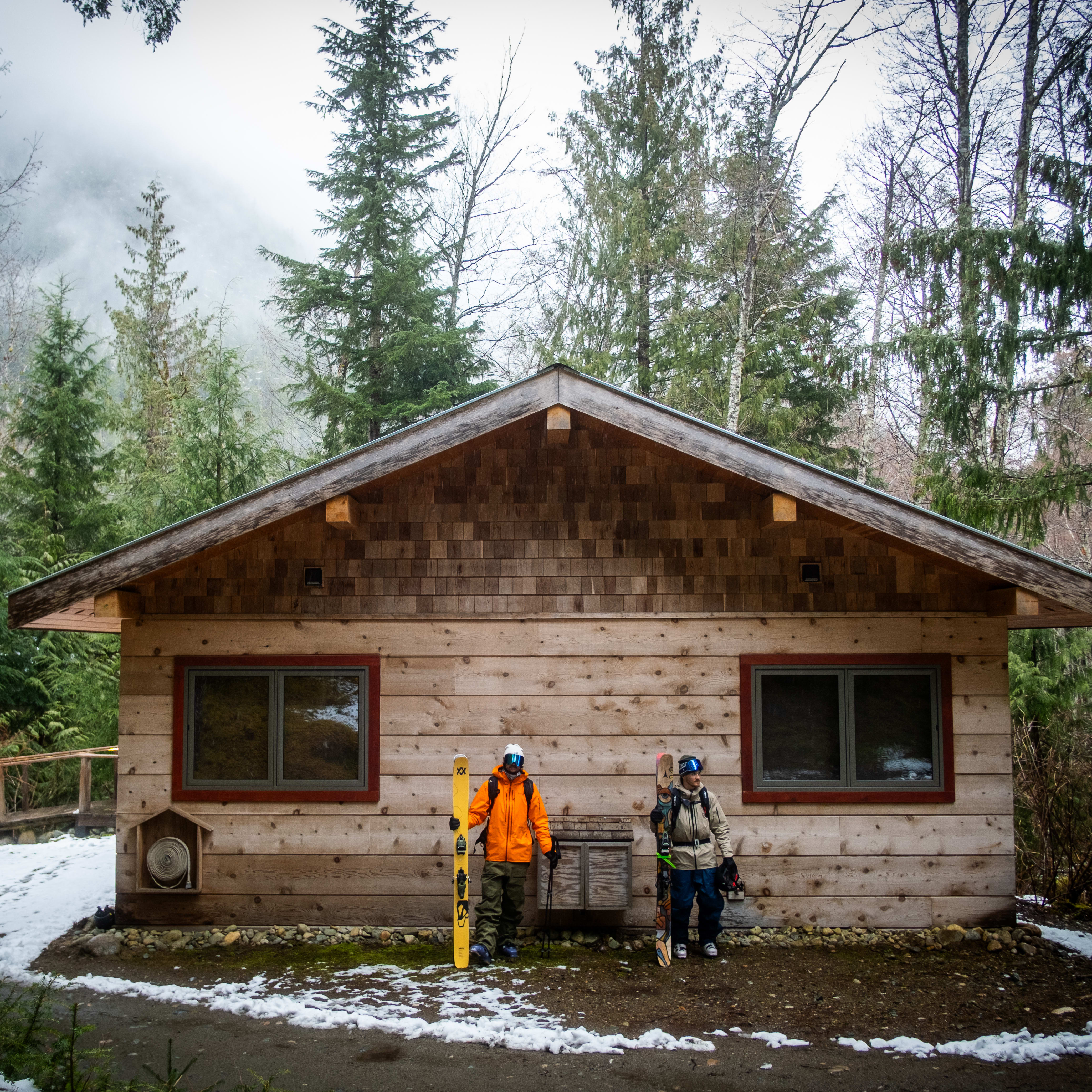 Two skiers in Norrøna gear stand outside a wood cabin in a forest. One wears an orange jacket.
