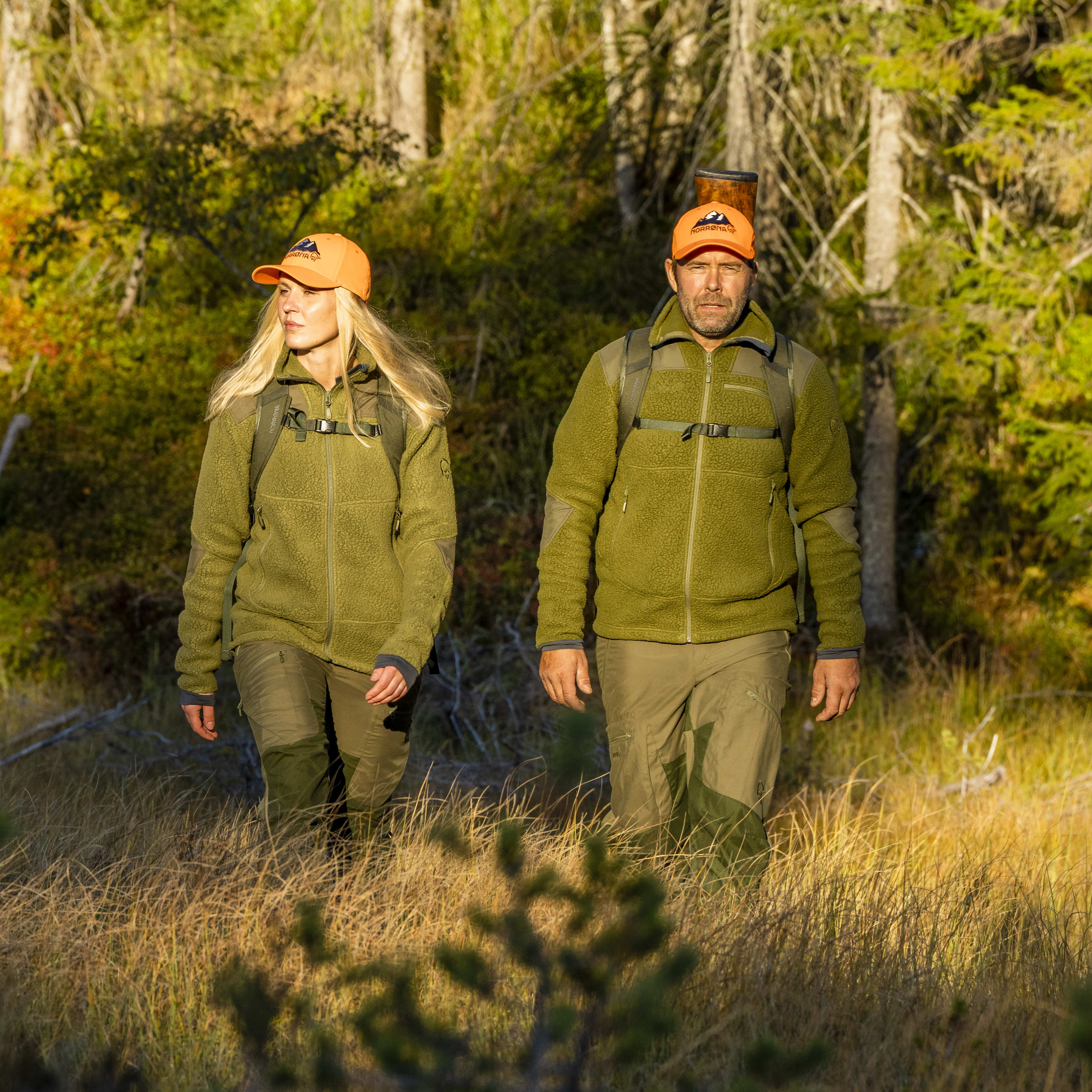 Two people wearing Norrøna clothing and orange hats walk through a grassy forest. 
