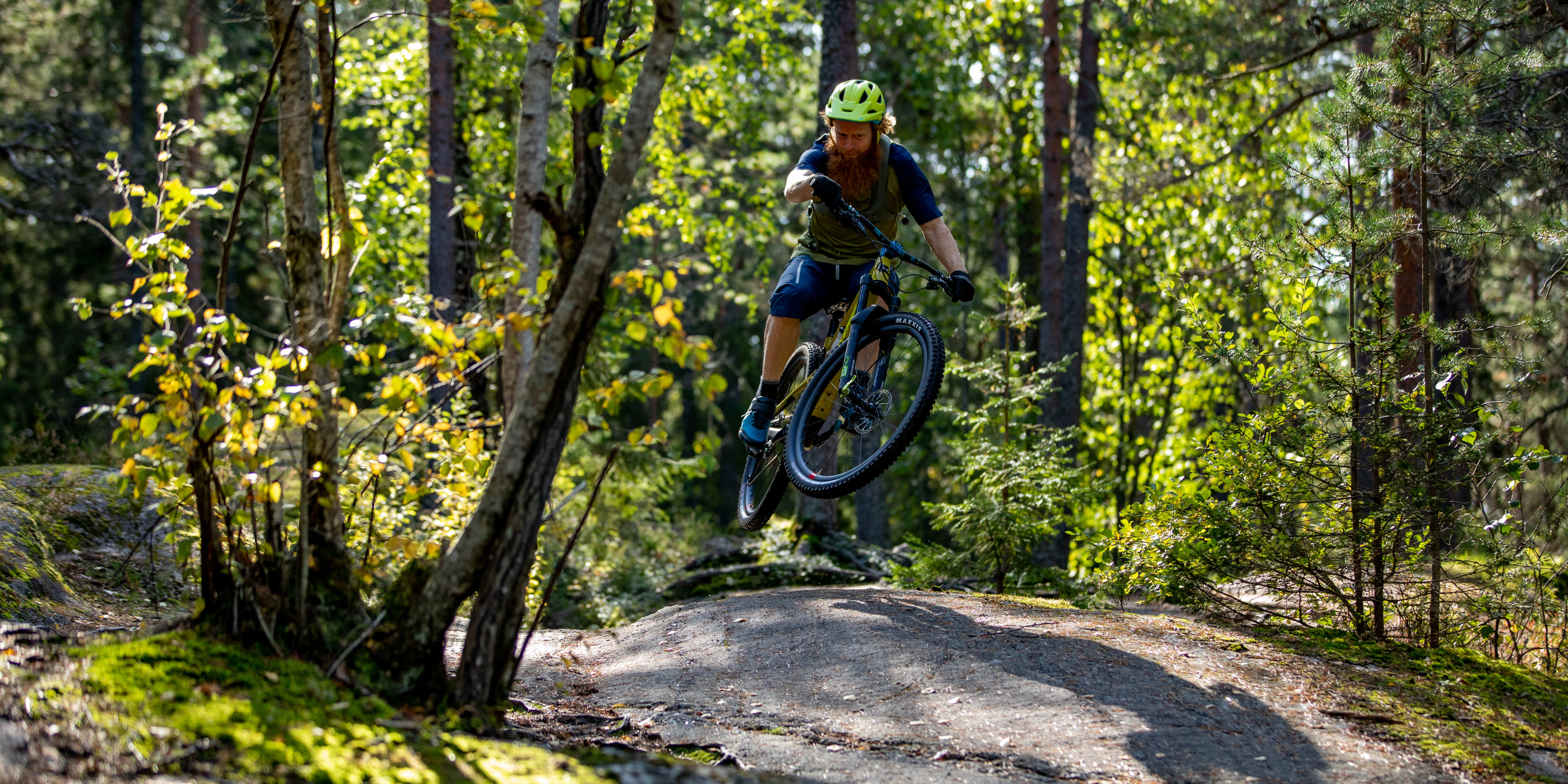 A man wearing a Norrøna outfit is riding a mountain bike in a forest. He is in mid-air, performing a trick over a rocky trail.  

