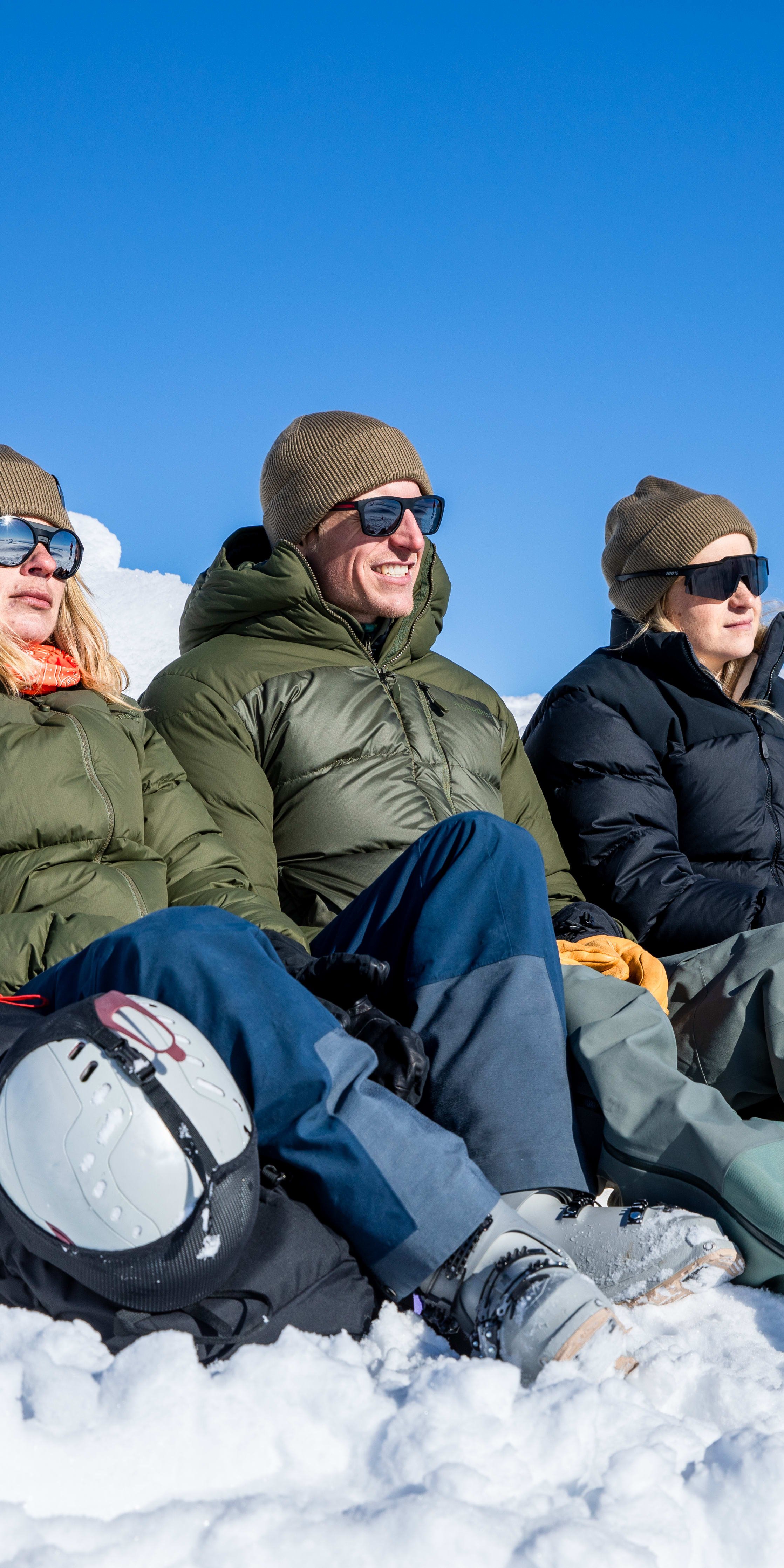 Three people wearing Norrøna gear and sunglasses sit in the snow with a clear blue sky above.  
