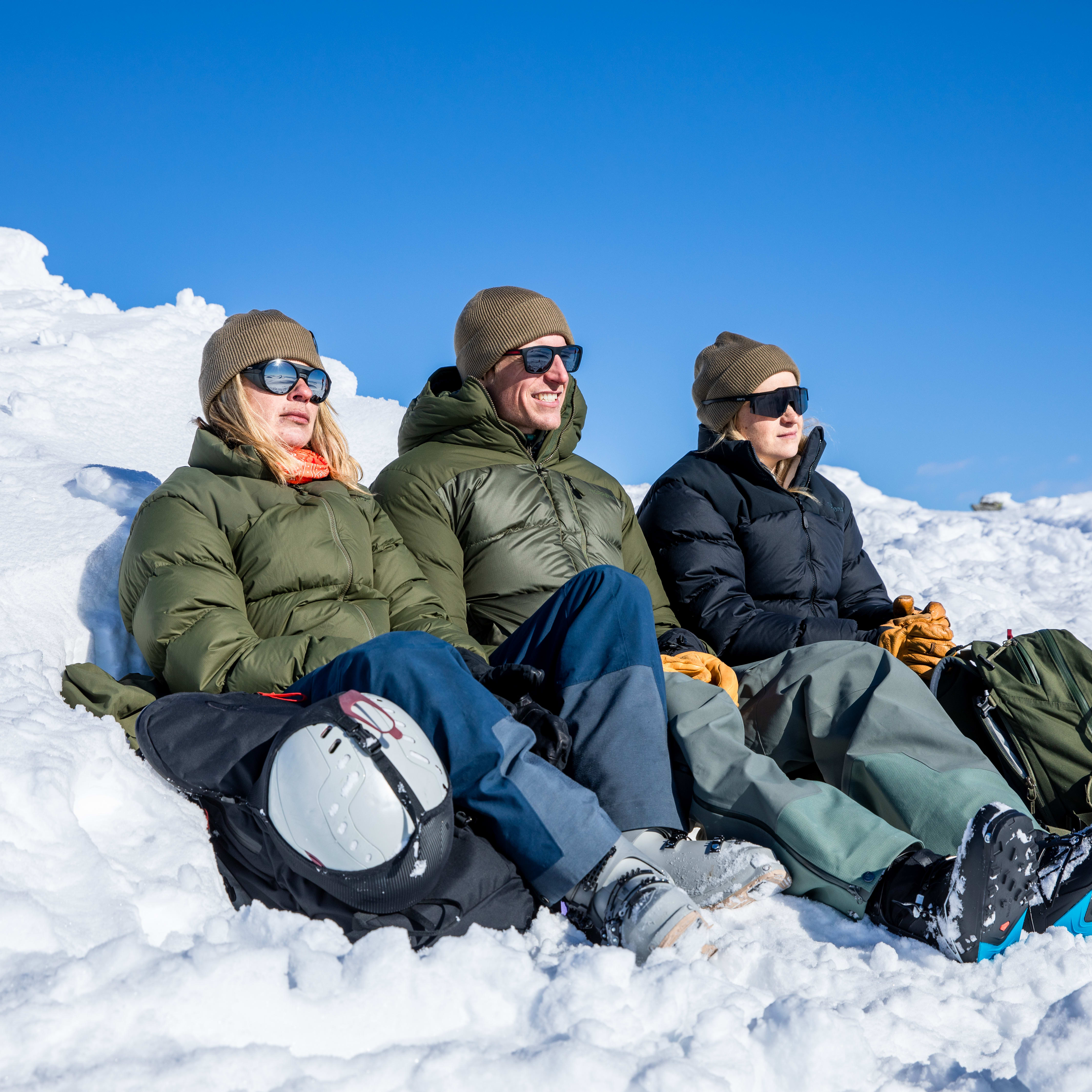 Three people wearing Norrøna gear and sunglasses sit in the snow with a clear blue sky above.  

