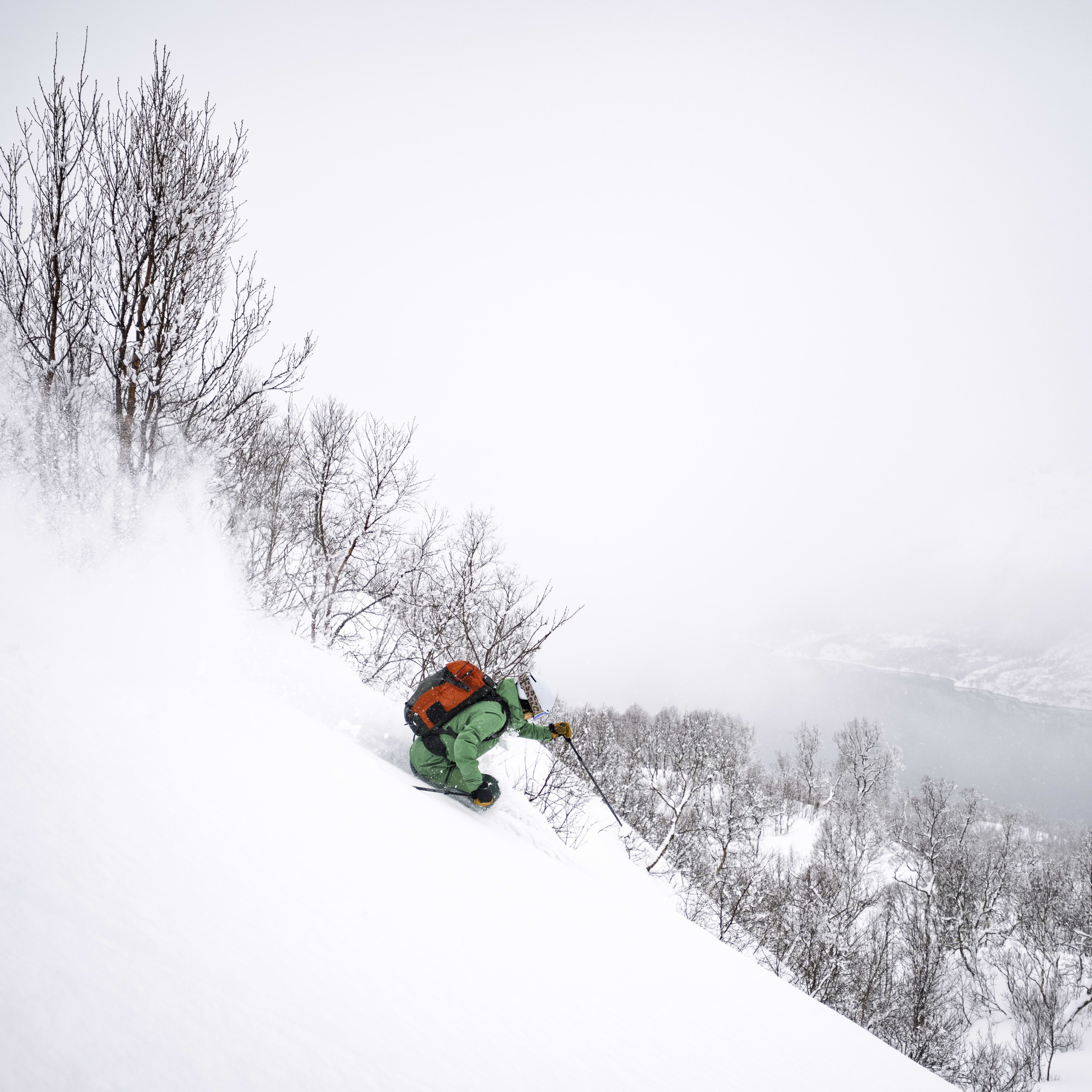 A person wearing a green Norrøna jacket and a backpack skis down a snowy slope with a lake and mountains in the background. 
