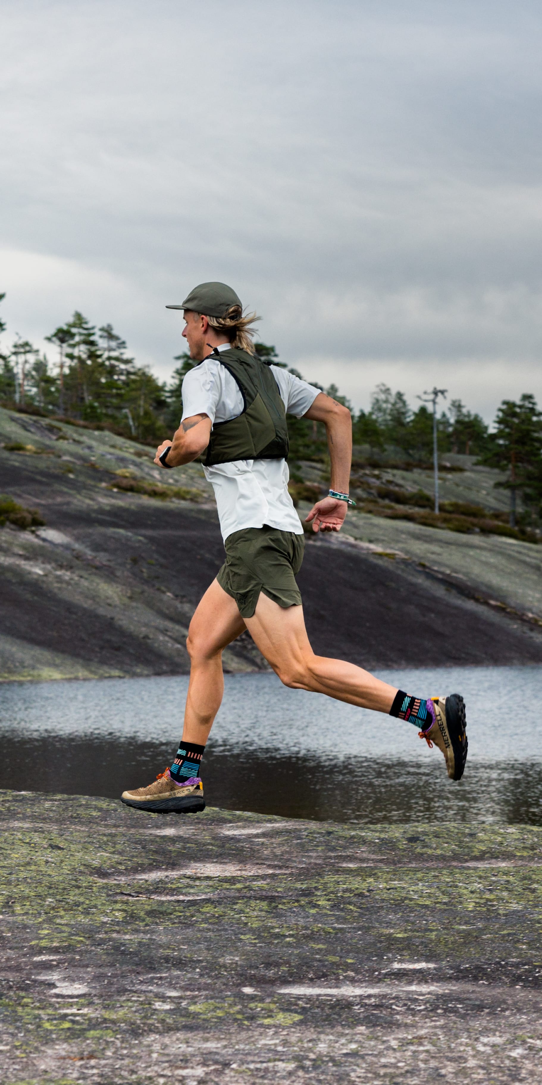 A man runs in Norrøna shorts and vest by a lake. He wears a hat, socks, and trail running shoes. Trees and gray rocks are in the background.
