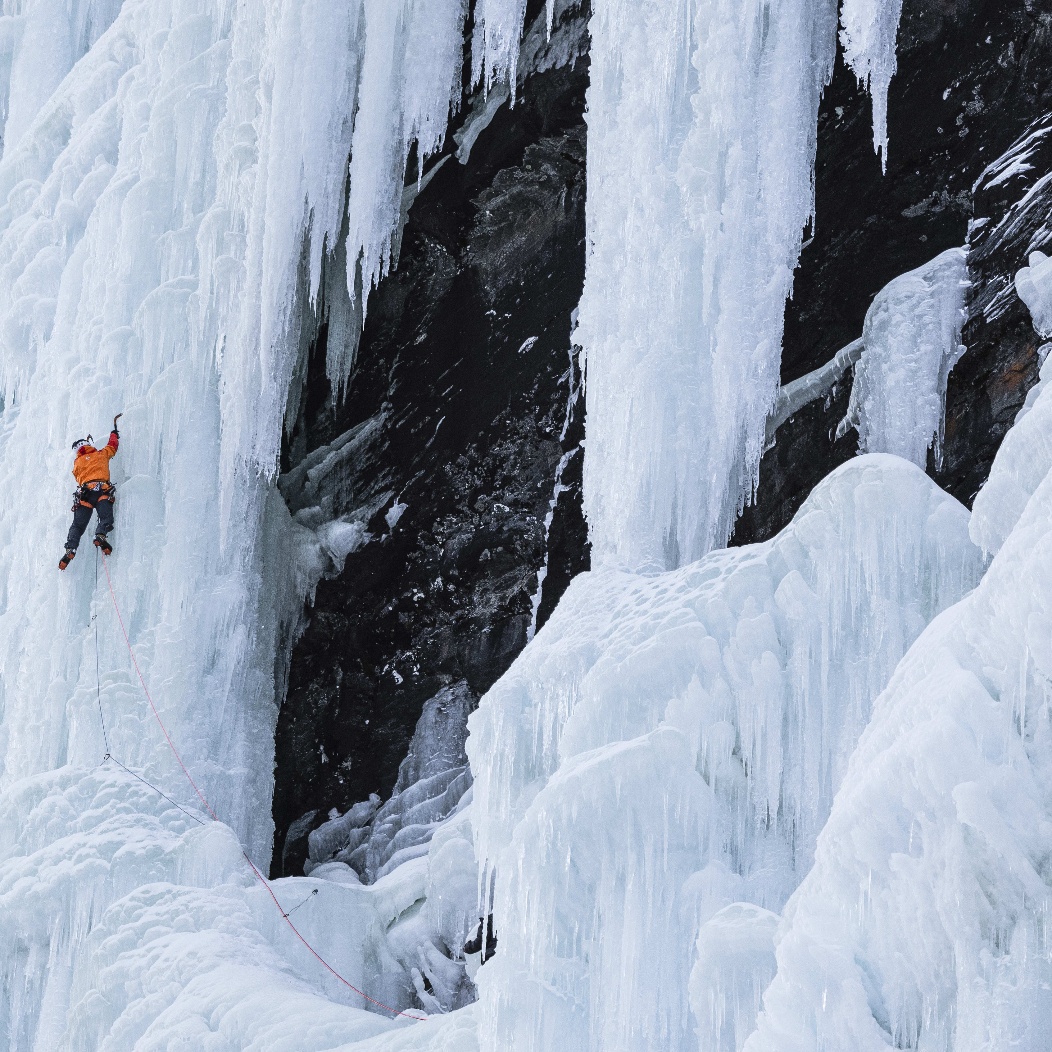 A person ice climbing a frozen waterfall, wearing an orange Norrøna jacket, with safety equipment. 
