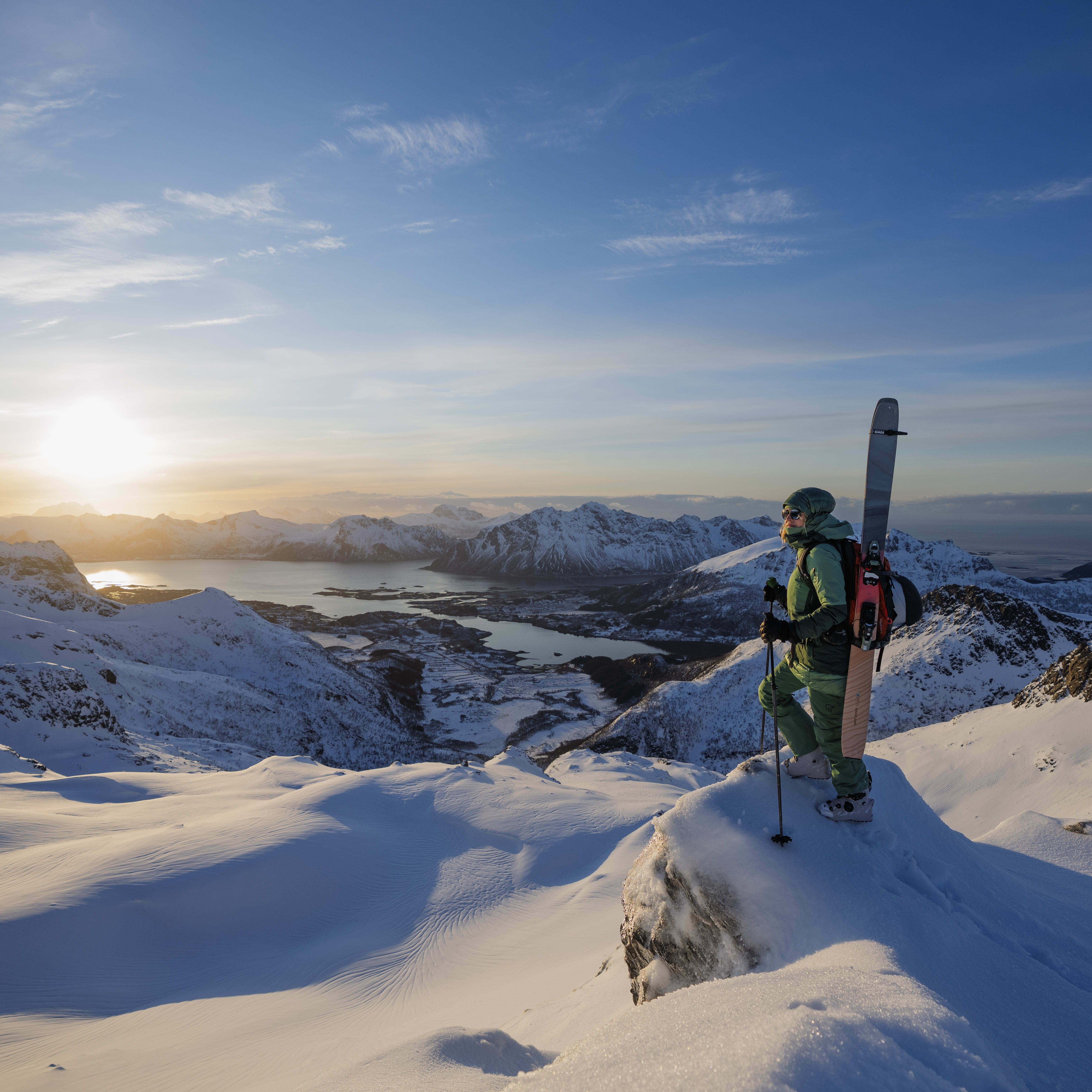 A skier stands on a snow-covered mountaintop, overlooking a stunning view of a fjord and mountains, bathed in the golden light of a sunset. They are wearing Norrøna gear. 
