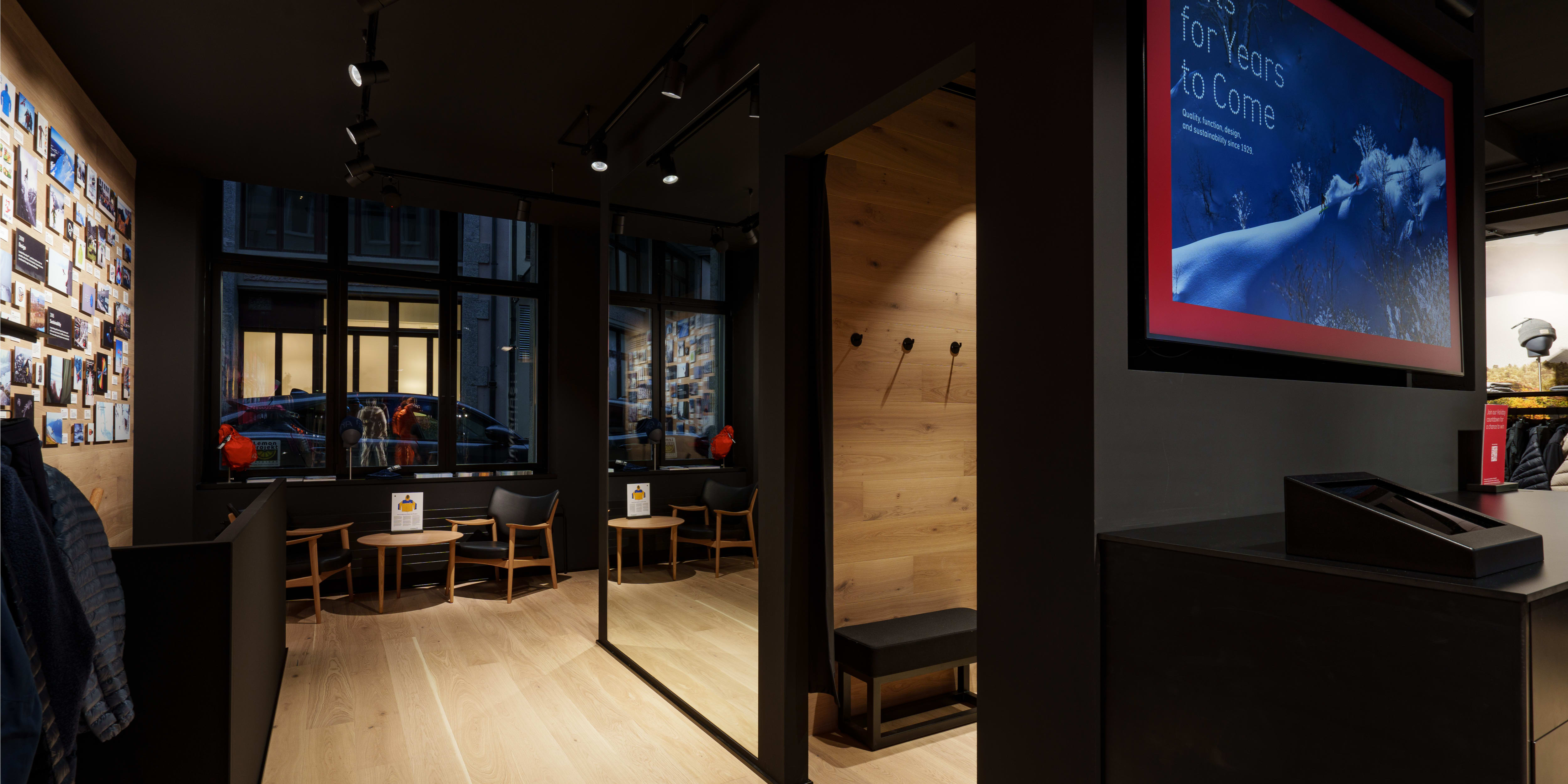 A Norrøna store interior with wood floors, a screen displaying winter sports advertising, and a black bench in the changing room. 
