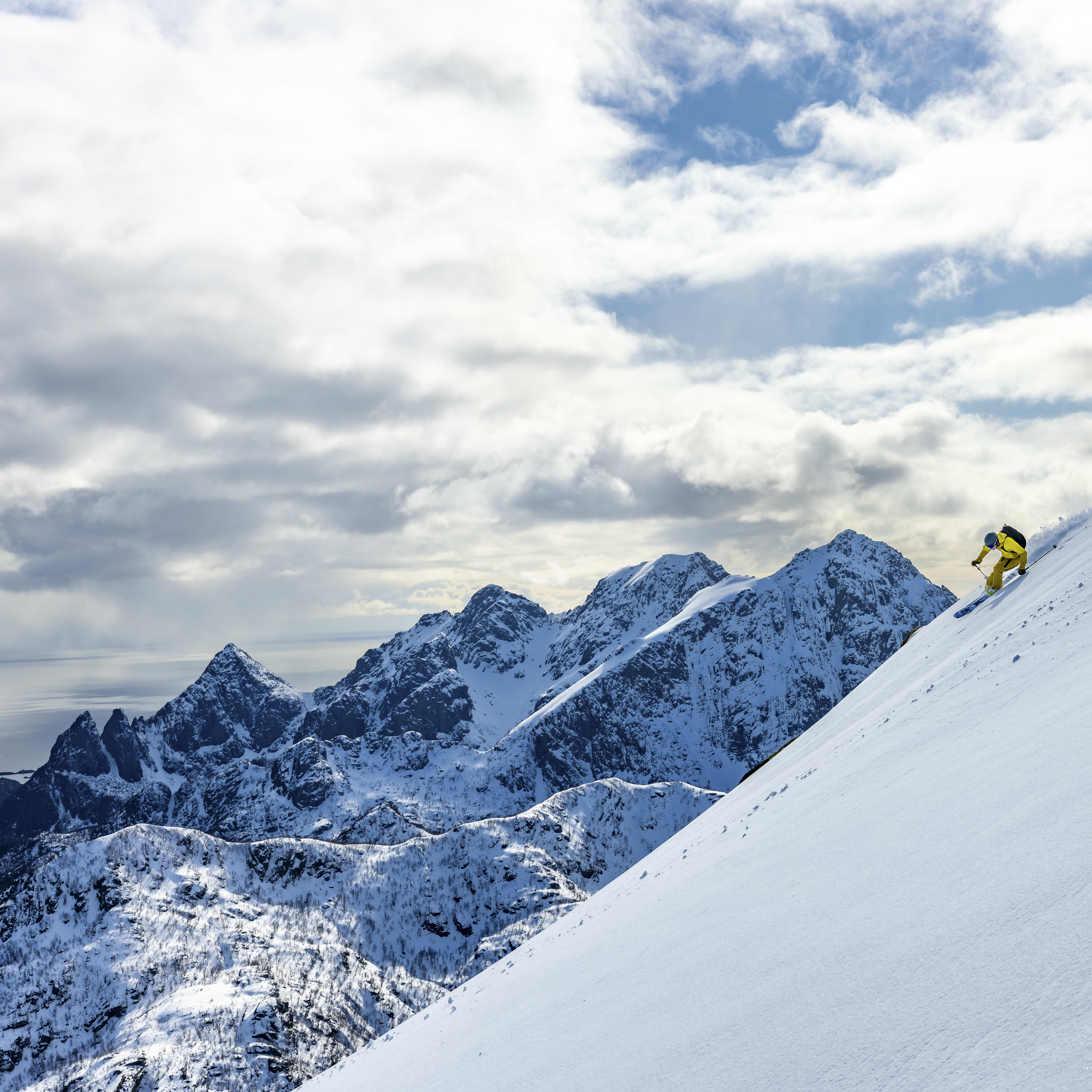 A skier in a Norrøna suit descends a snowy mountain slope against a backdrop of a snow-covered mountain range and a blue sky with white clouds.