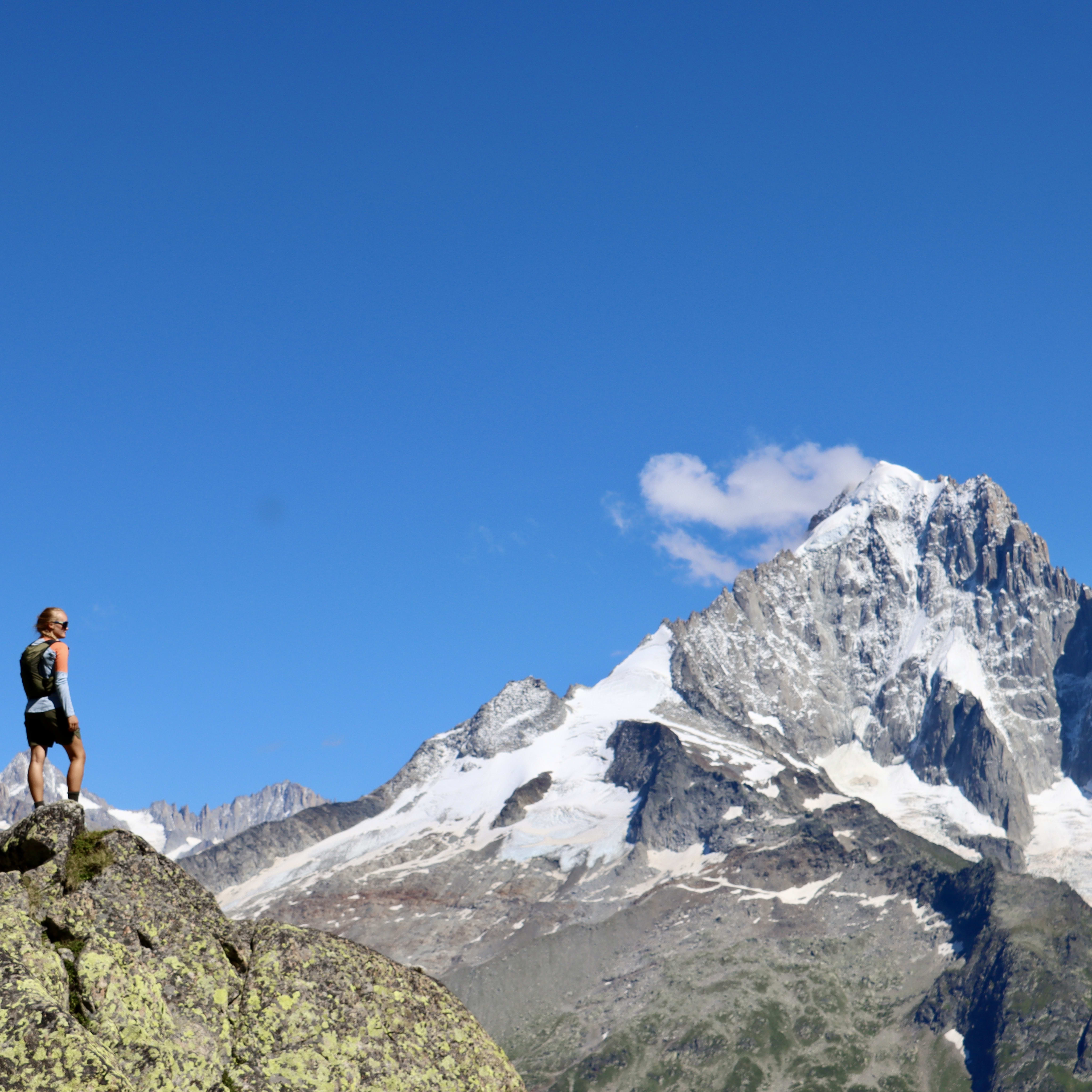 A person wearing Norrøna clothes stands atop a rock with mountain ranges in the background under a clear blue sky.