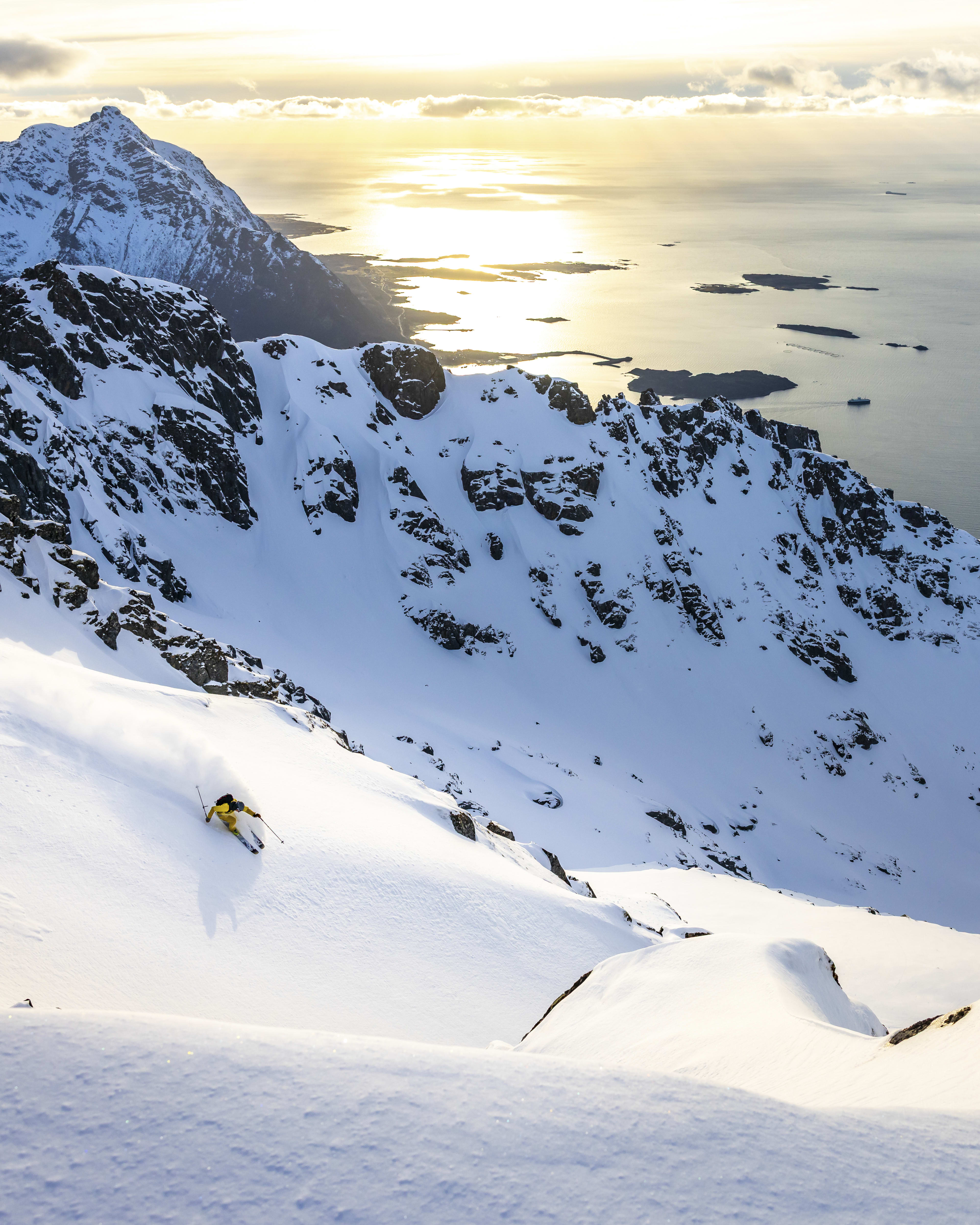 A skier in yellow clothing descends a snow-covered mountainside. The mountains overlook a vast, blue ocean in the distance. 

