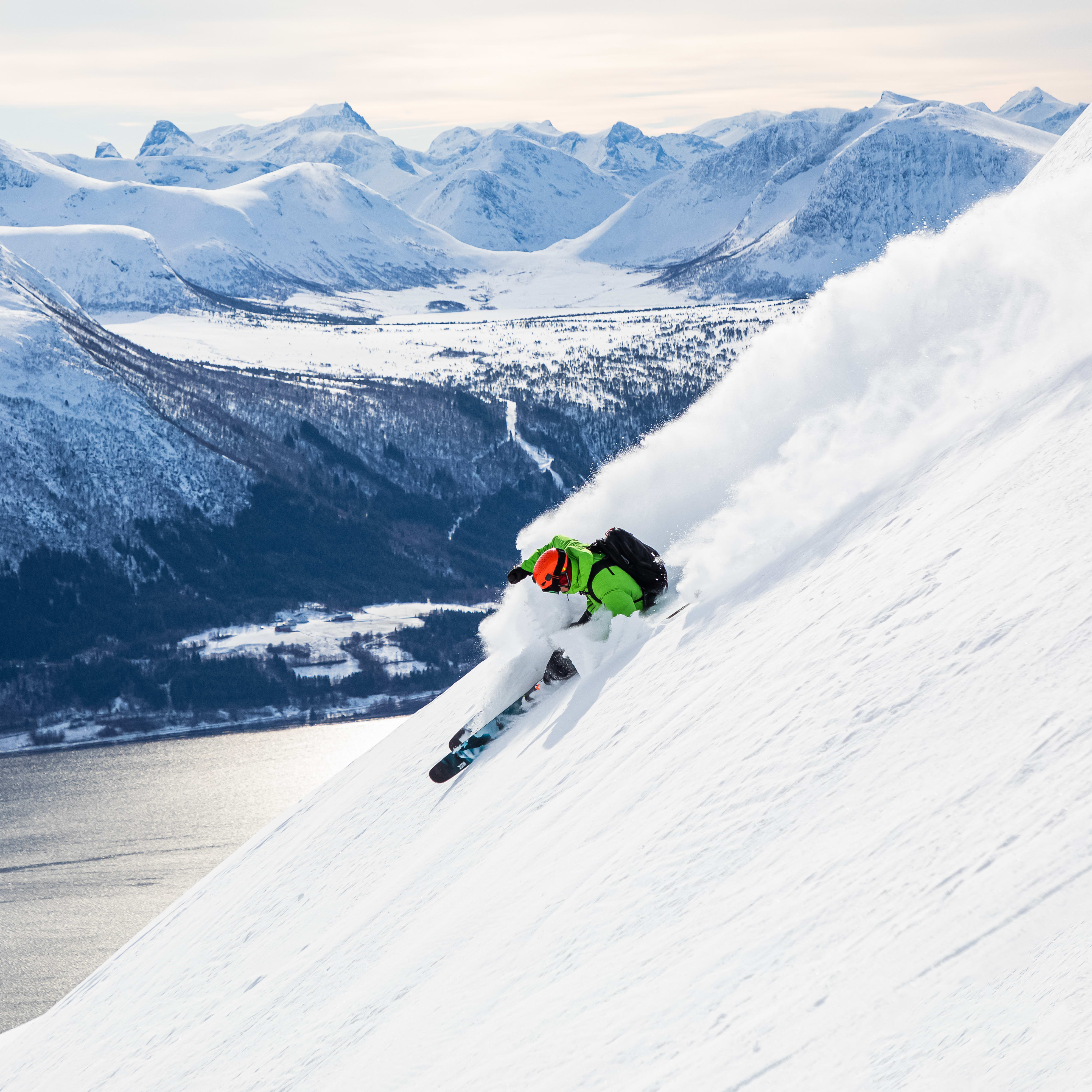 A skier in green Norrøna clothing carving a turn through fresh powder on a snowy mountainside.
