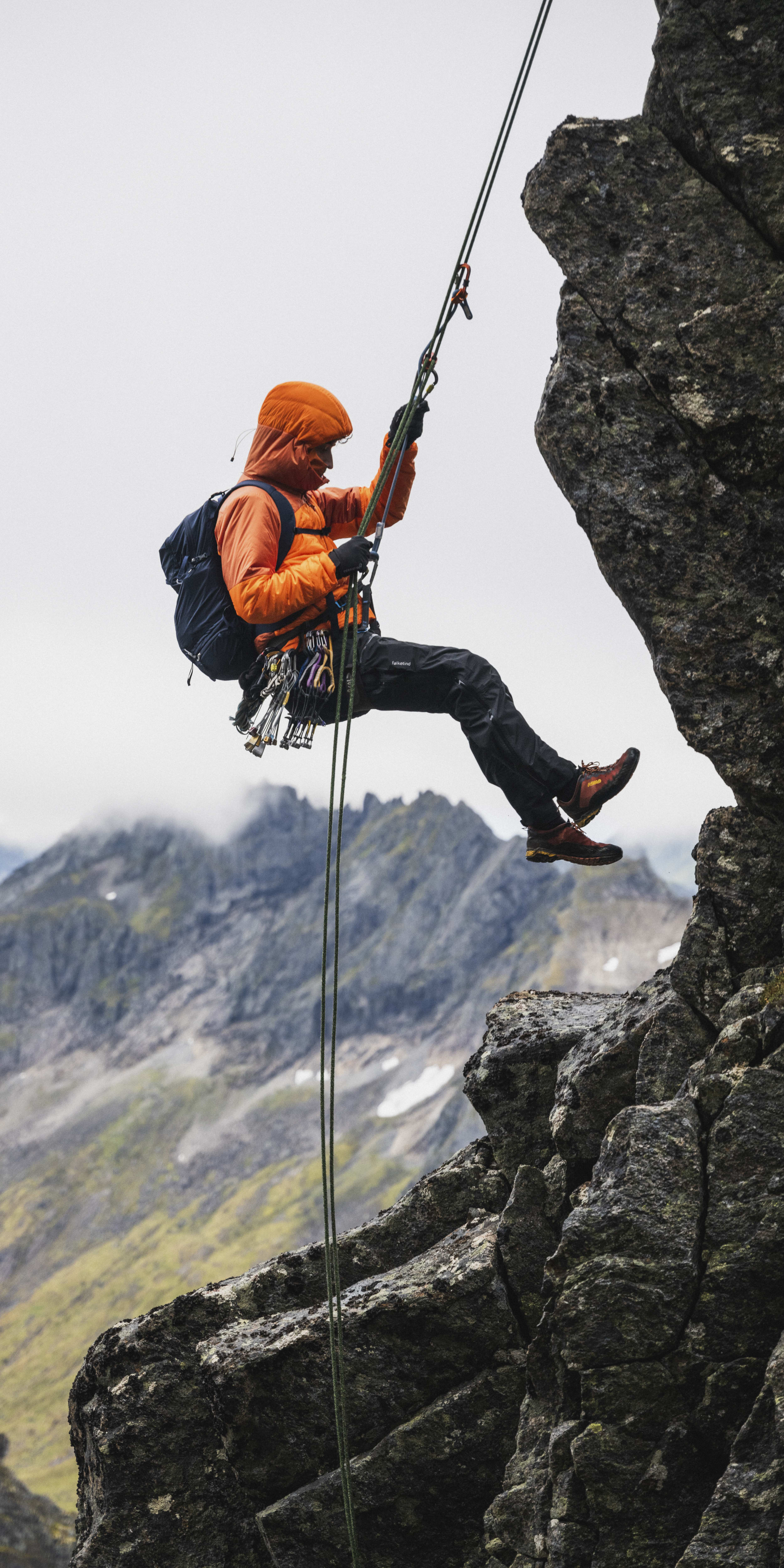 A man wearing an orange Norrøna jacket rappels down a rocky cliff face.