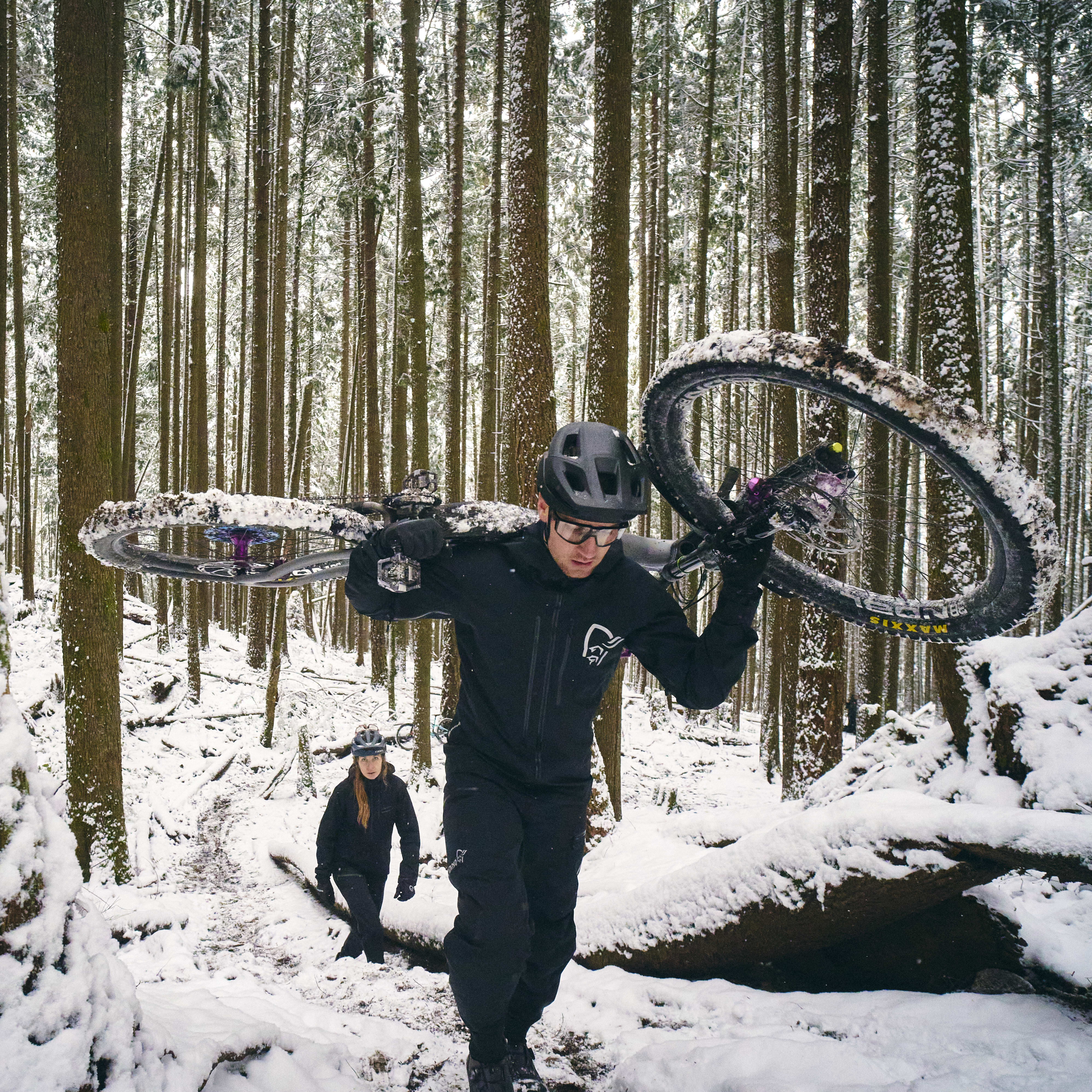 A man in a black Norrøna jacket carrying a mountain bike through a snowy forest. A woman in a black jacket walks behind him.