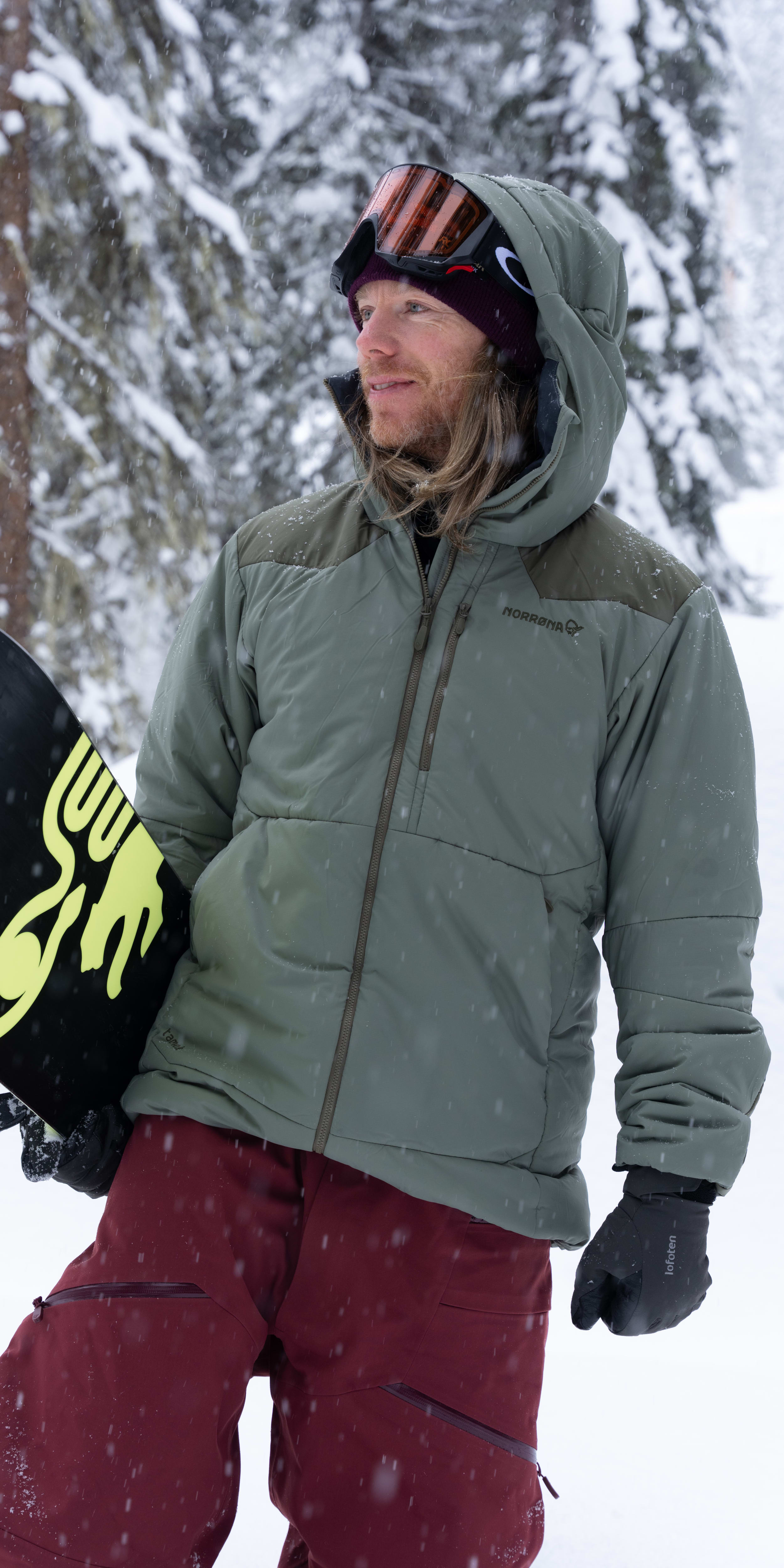 A snowboarder in a green Norrøna jacket and red pants holds a snowboard in a snowy forest. He wears goggles and gloves. It is snowing.