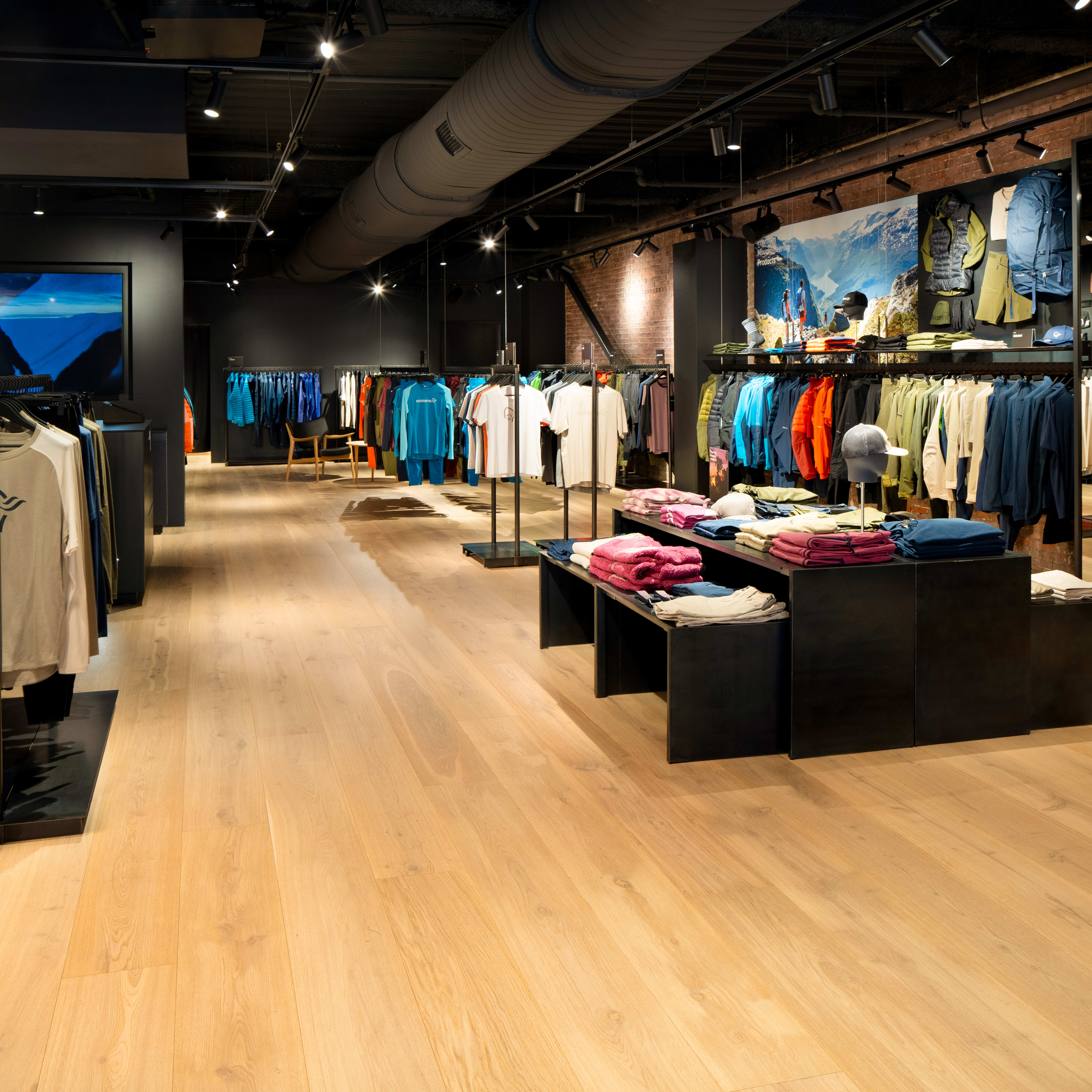 A Norrøna store with mannequins displaying clothing, a rack with Norrøna apparel, and a table with folded clothing.  The store has a wooden floor, brick wall and black metal fixtures. 
