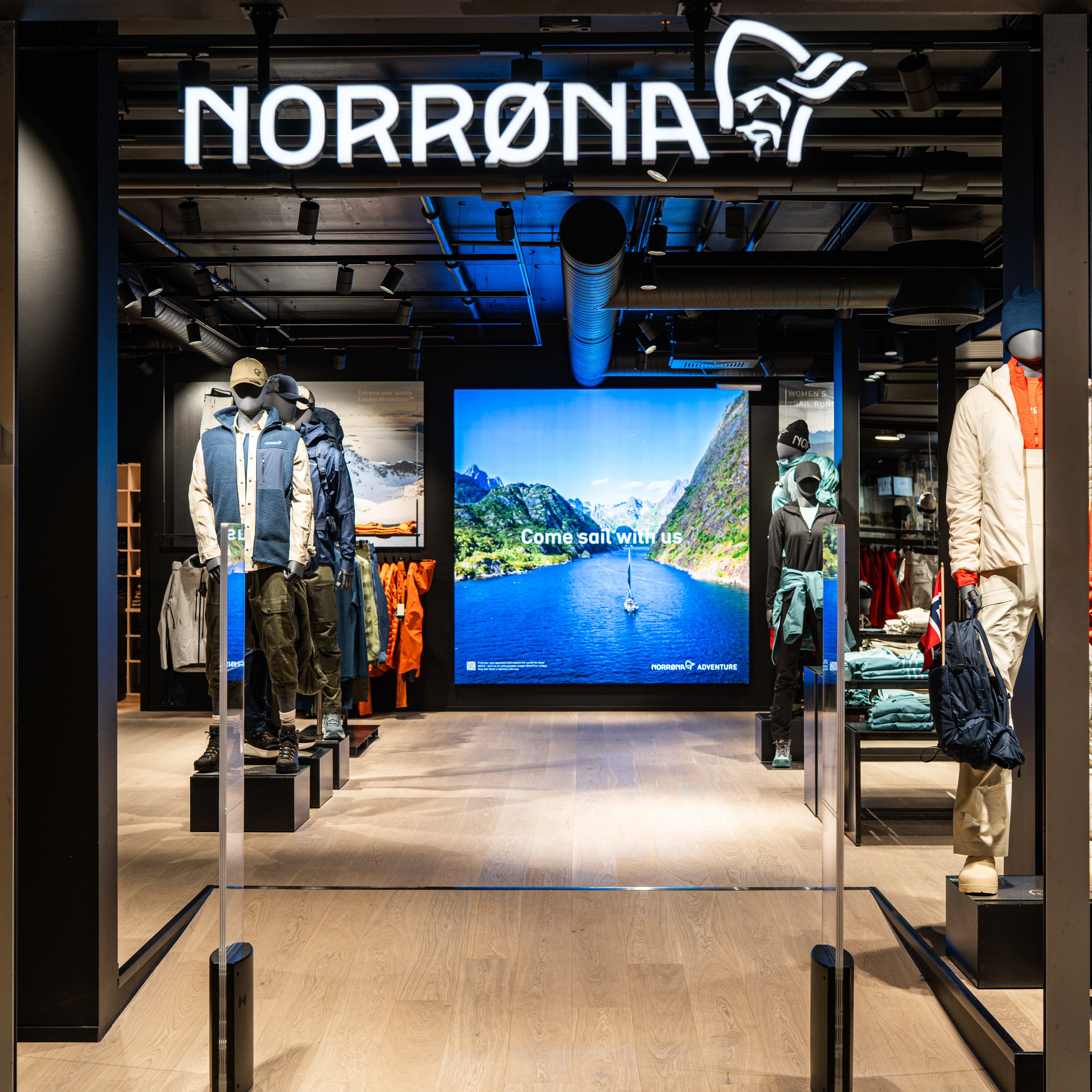Storefront of a Norrøna store with mannequins displaying outdoor clothing, and a screen showing a Norwegian fjord.