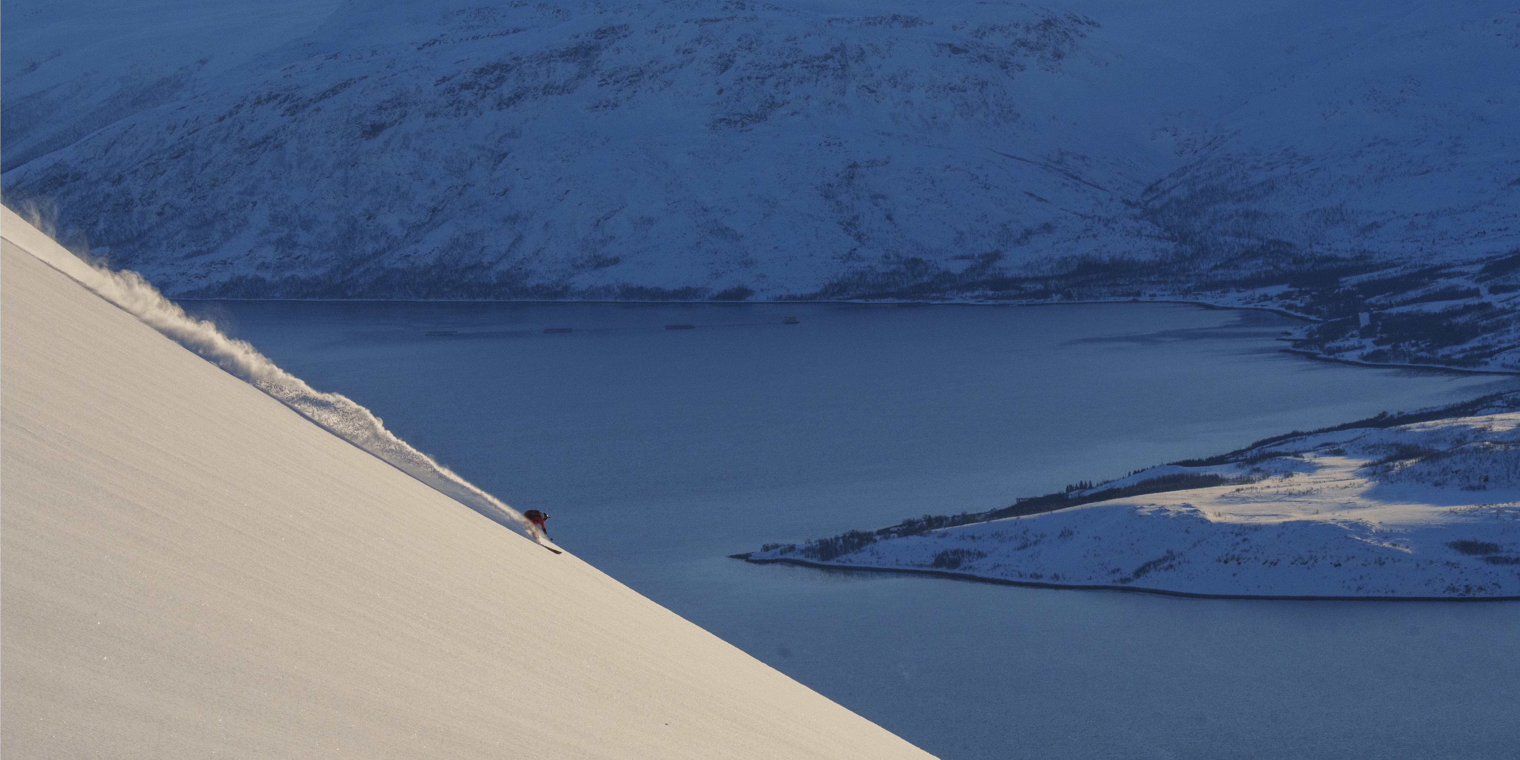 A skier in a Norrøna jacket descends a snowy slope, kicking up snow. A blue fjord and mountains are in the background.

