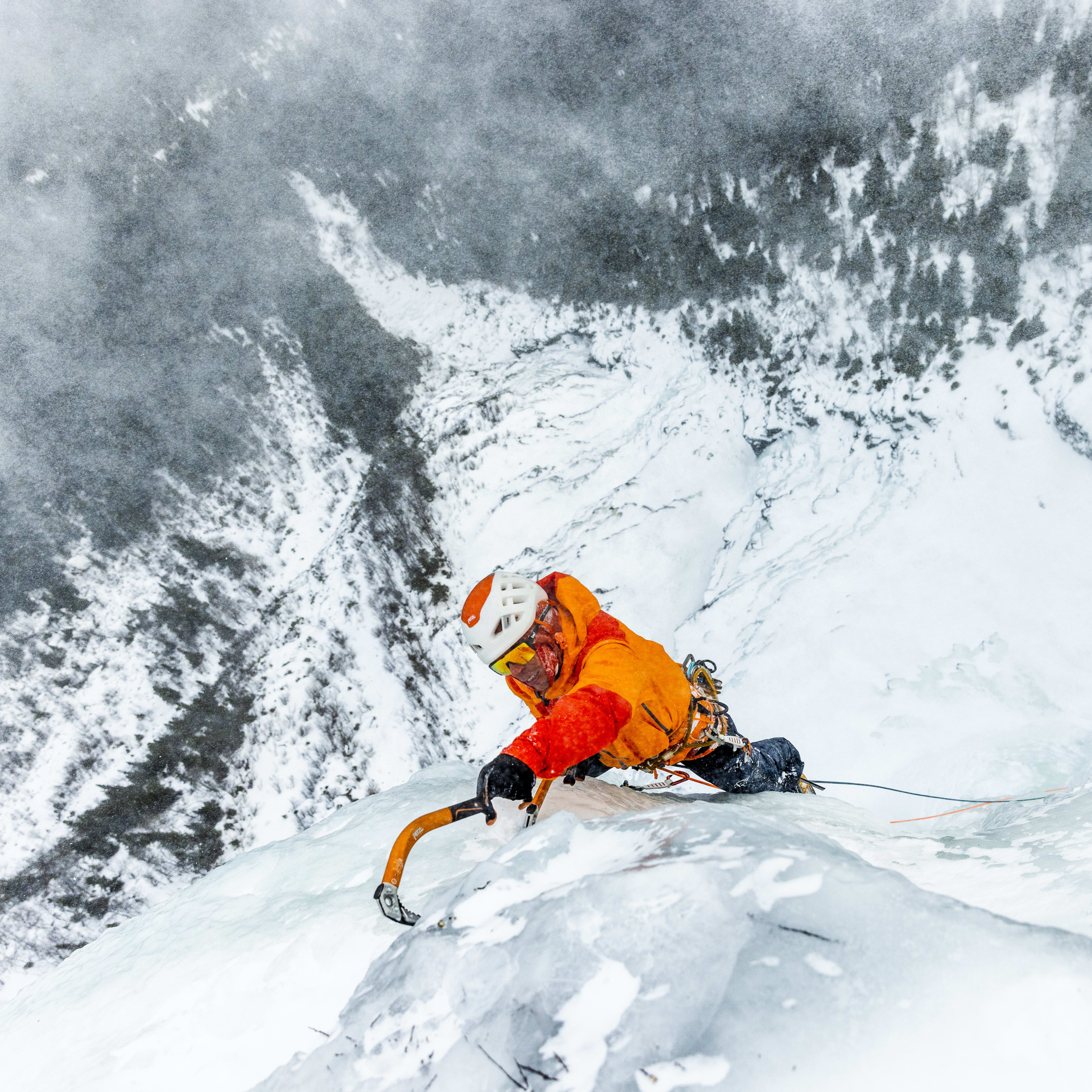 Matthias Scherer ice climbing Hydnefossen in an orange Norrøna jacket and helmet in Hemsedal. He is wearing Norrøna trollveggen Gore-Tex Pro Light jacket and pants. Picture is taken from above the climber, looking down into the ice cold valley.