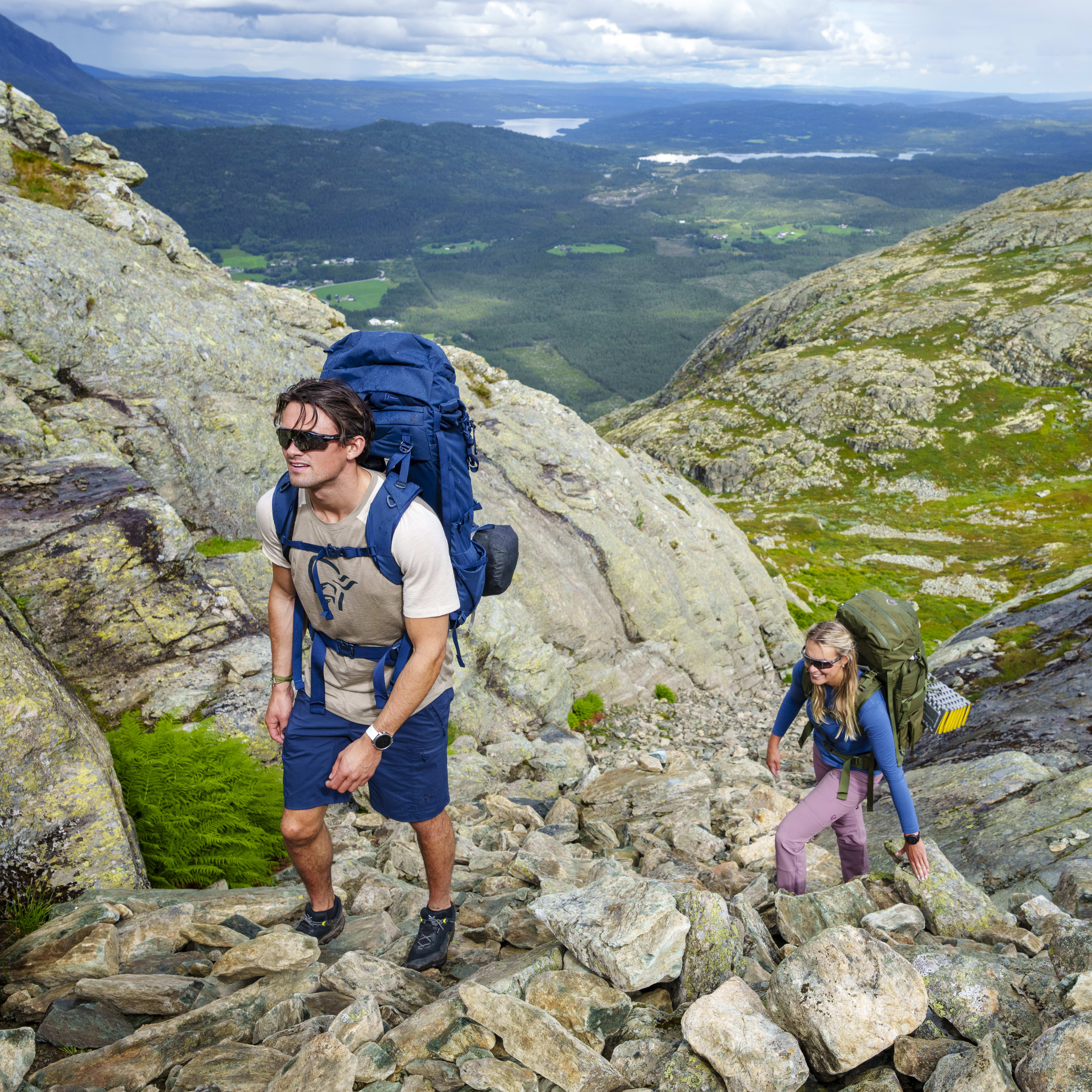 Two people wearing Norrøna gear hiking on a rocky mountain path.  The path goes up to a mountain range and a lake.  
