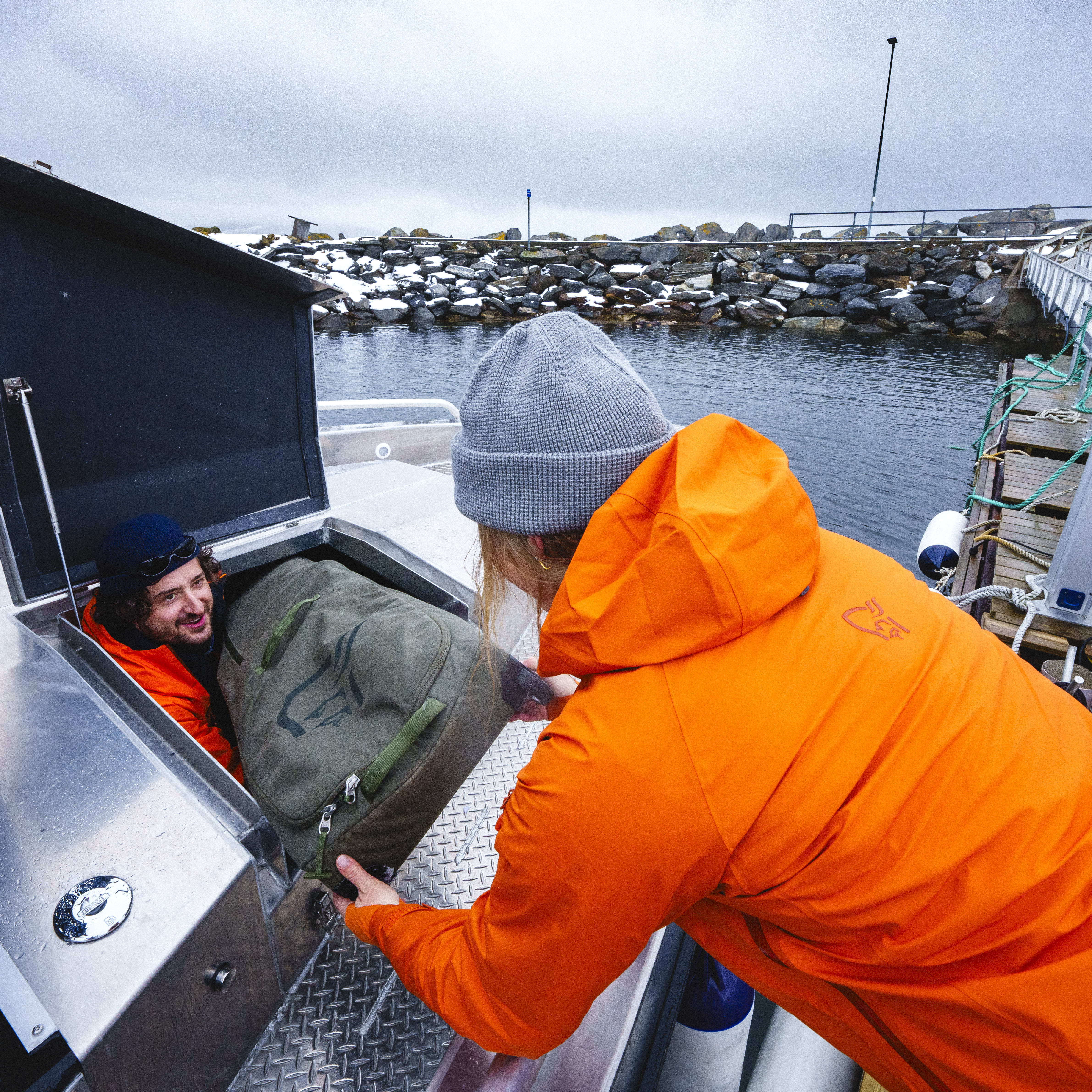 A person wearing an orange Norrøna jacket and a grey beanie is putting a large green Norrøna bag into the open storage compartment of a boat. Another person is inside the compartment, looking up.