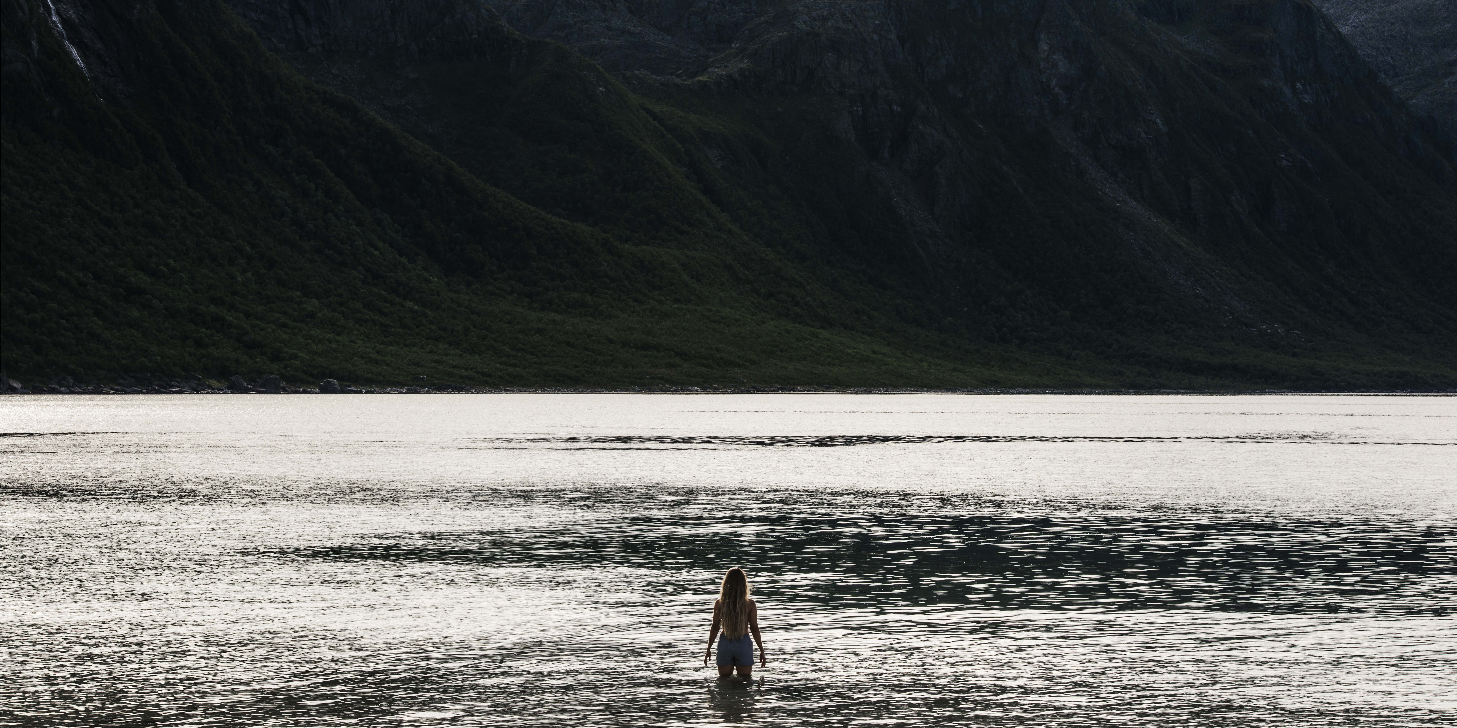 A woman wearing a Norrøna shirt stands in a body of water facing a mountain range.