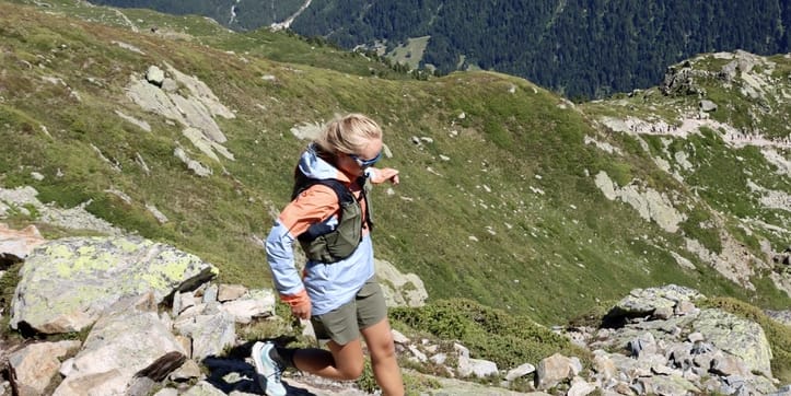 A woman hikes downhill on a rocky trail, wearing a Norrøna jacket and vest. Majestic, snow-capped mountains rise in the background.