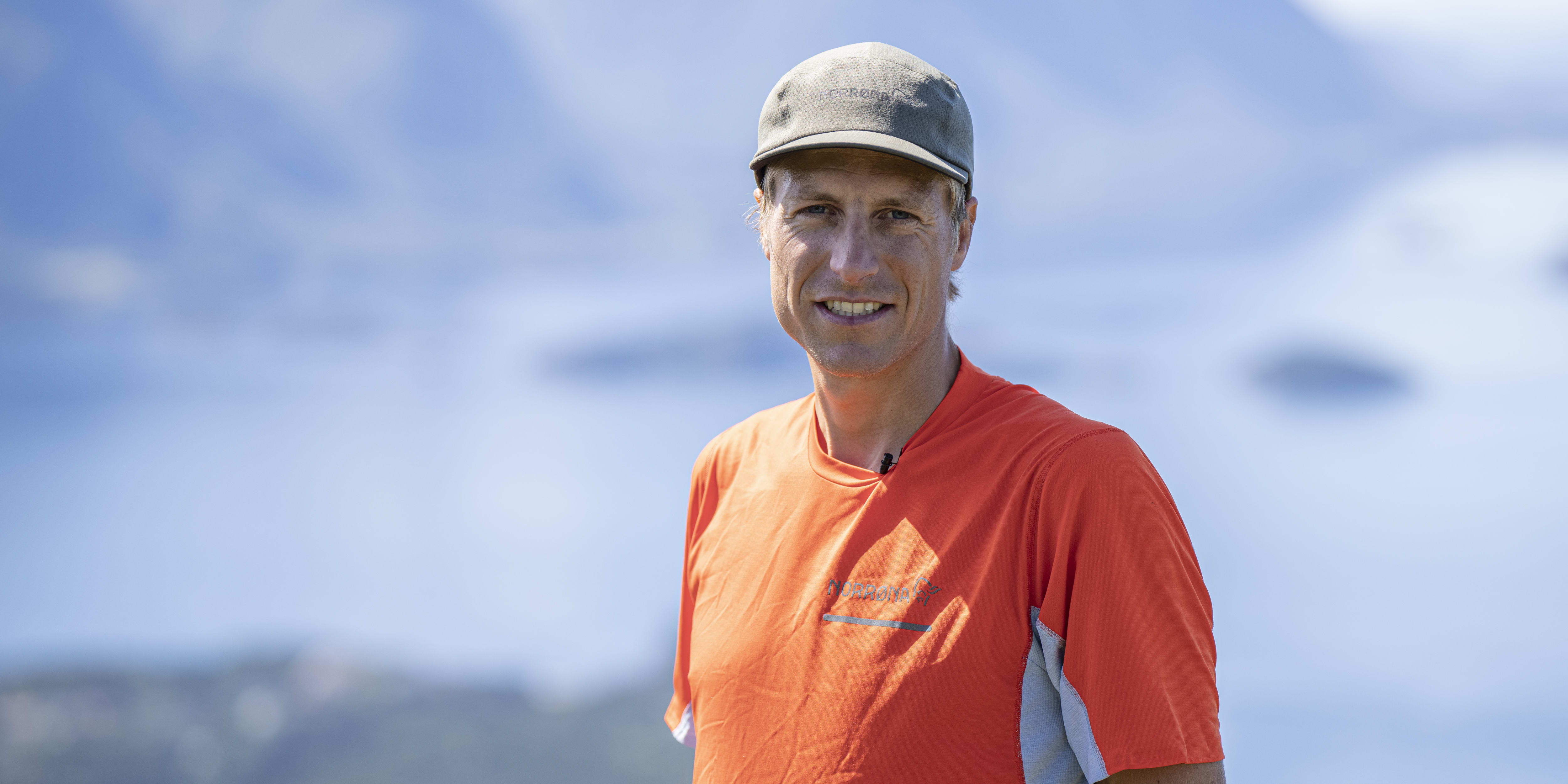 A man smiling at the camera. He's wearing a Norrøna shirt and a hat, standing in front of a blurred mountain background. 
