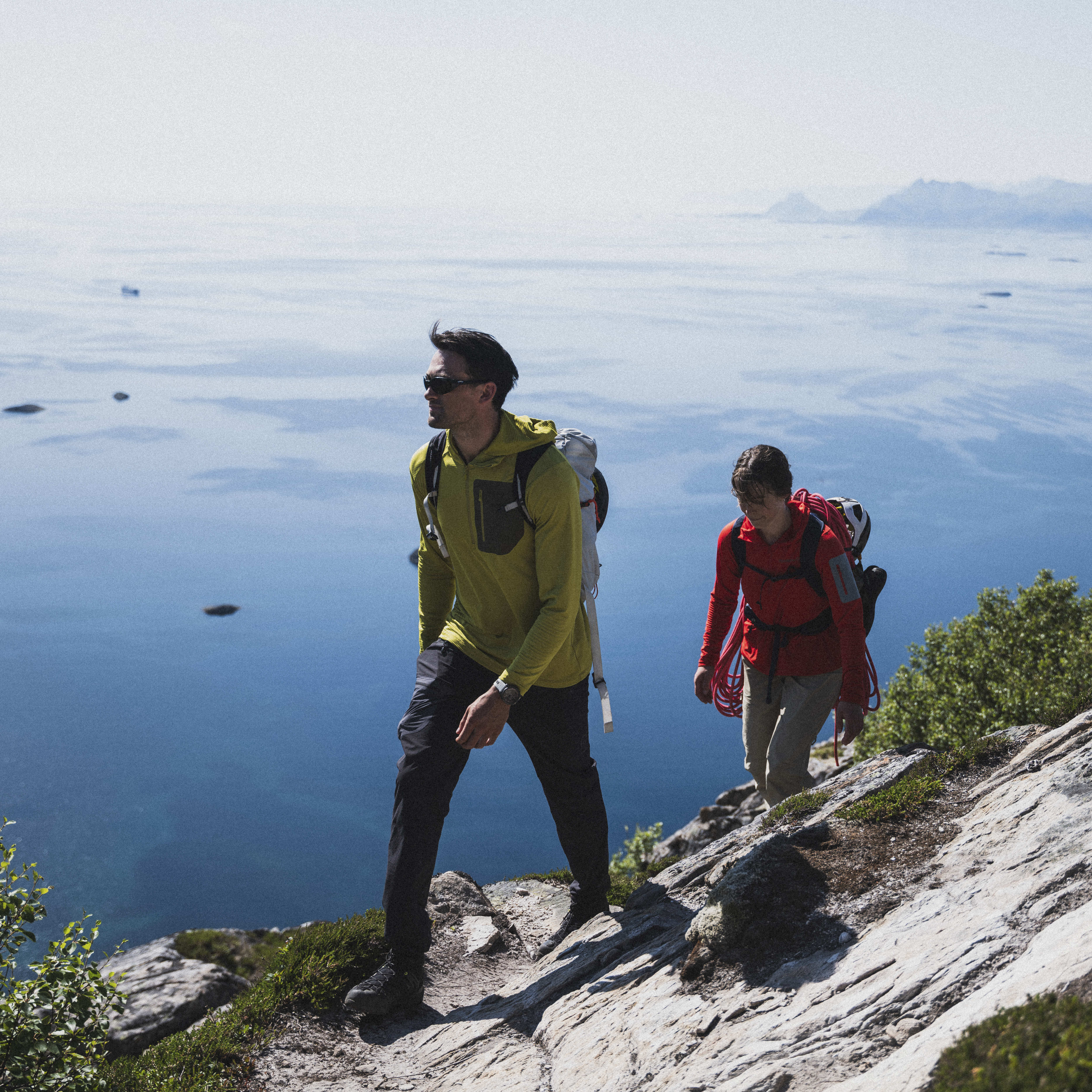 Two hikers on a rocky trail with a vast ocean view. The man wears a yellow Norrøna hooded top and sunglasses. The woman wears a red Norrøna top and carries climbing gear.
