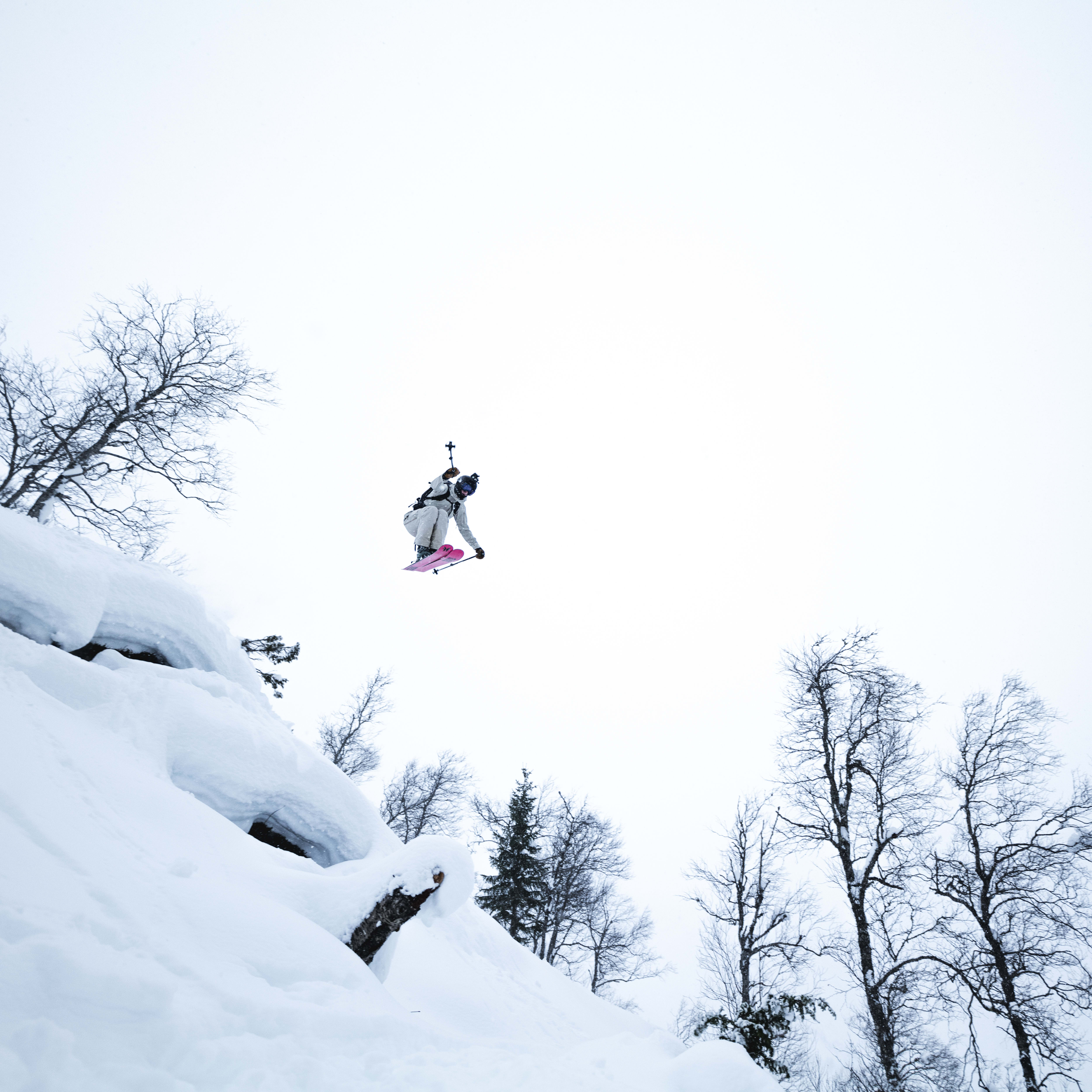 A person in a white ski suit and a black helmet is mid-air while skiing, with two black ski poles and pink skis. They are in the air above snow-covered terrain and bare trees. The sky is clear and light blue.